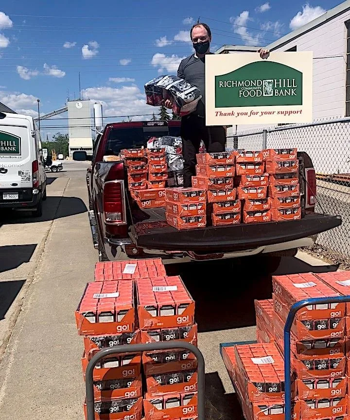 Man wearing a black face mask holding boxes, standing in the truck bed filled with boxed food donations, at a food bank parking lot.