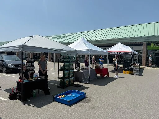 Outdoor market with multiple vendor tents and tables in a parking lot, people browsing, and a building with a green roof in the background.