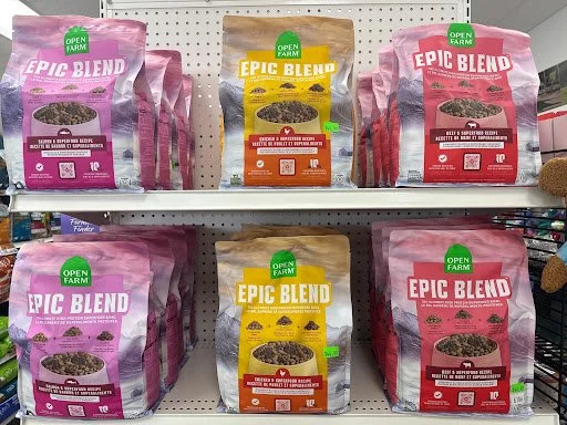 Six bags of Open Farm Epic Blend dog food on store shelves, with three pink and three yellow bags.