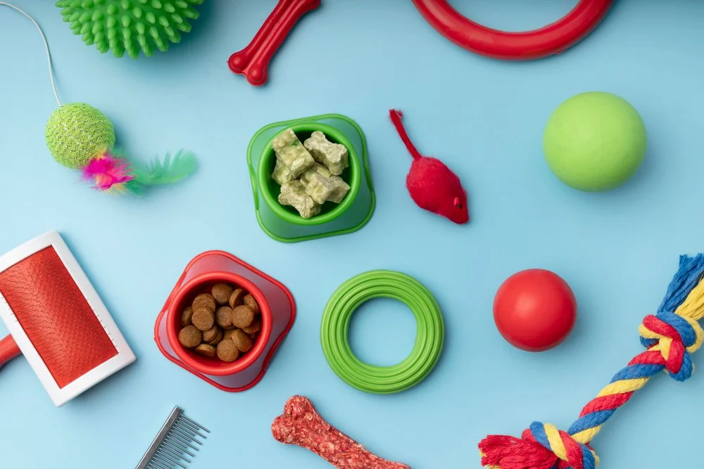 Various colorful dog toys and treats on a light blue surface, including squeaky toys, chew toys, a ball, a rope, and snacks.