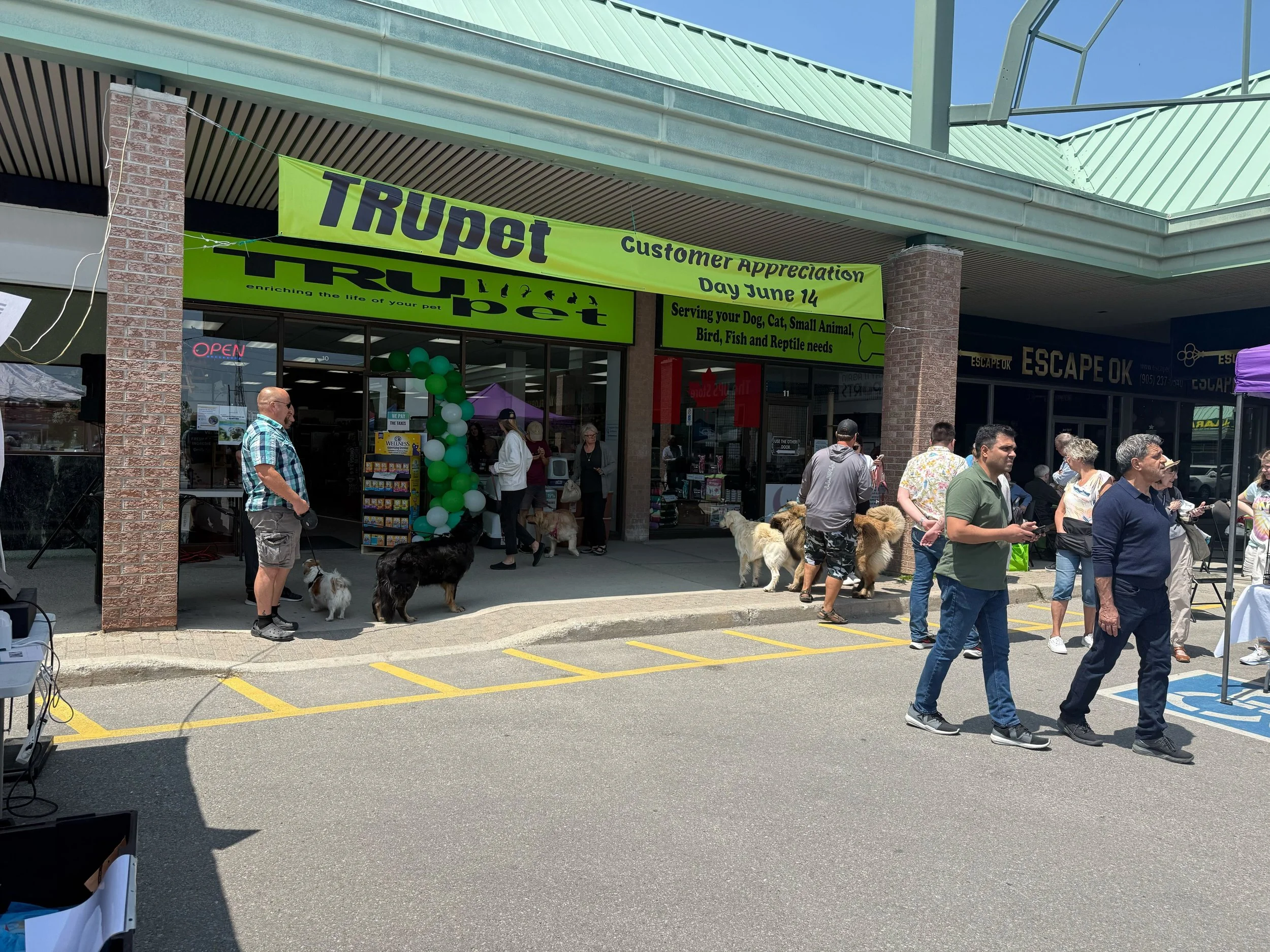 People with dogs gathered outside a pet store during a customer appreciation event, with a large green banner and balloons.