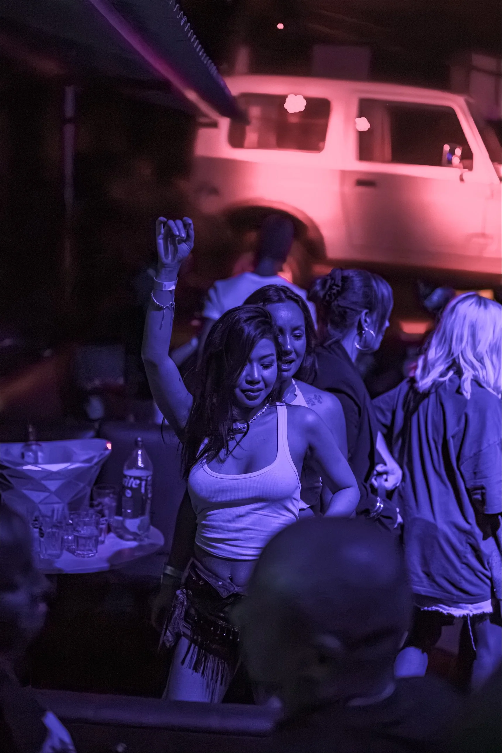 Menschen tanzen in einer dunklen, neonlichtdurchfluteten Partyumgebung, eine Frau in weißem Tanktop und Fransenrock im Mittelpunkt, im Hintergrund eine pink beleuchtete alte Mercedes-Benz.