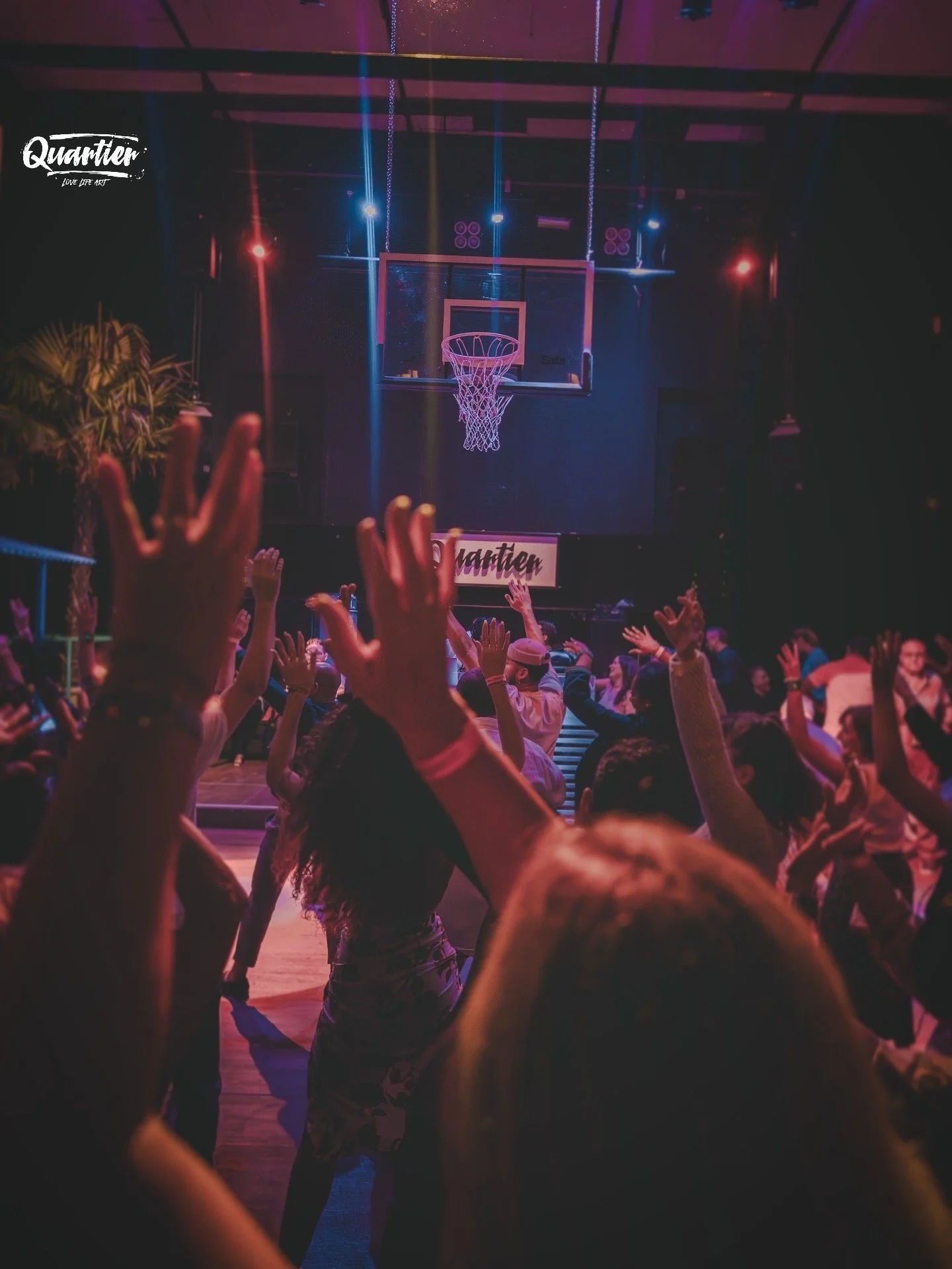 Menschen tanzen in einer Diskothek oder einem Veranstaltungssaal mit roten, blauen und lila Lichter, während sie den Händen in die Luft halten. Im Hintergrund befindet sich eine Basketball-Korb mit einer LED-Beleuchtung. Es gibt eine Bühne mit einem Schild, auf dem 'Quarter' steht.