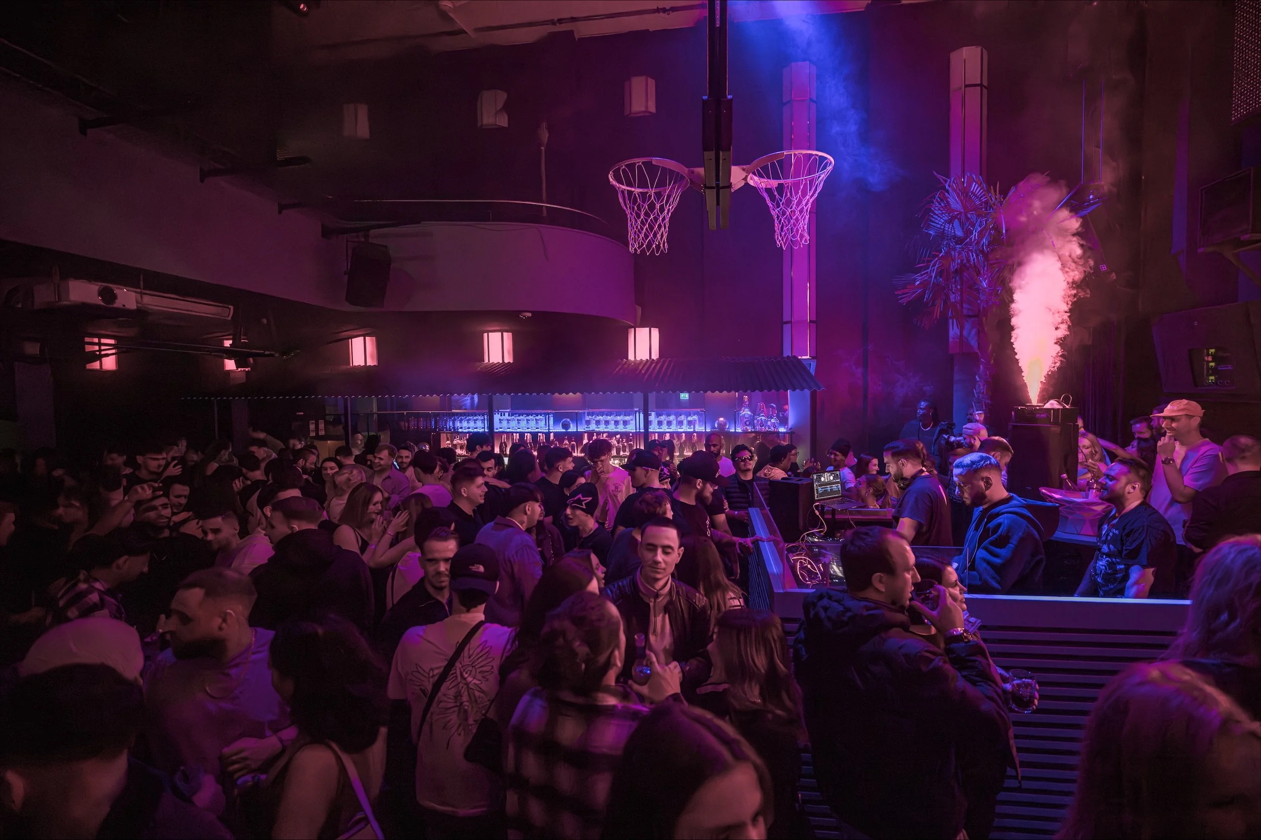 Menschen tanzen und feiern in einer Diskothek mit buntem Lichteffekten und DJ-Setup, im Hintergrund eine Bar.