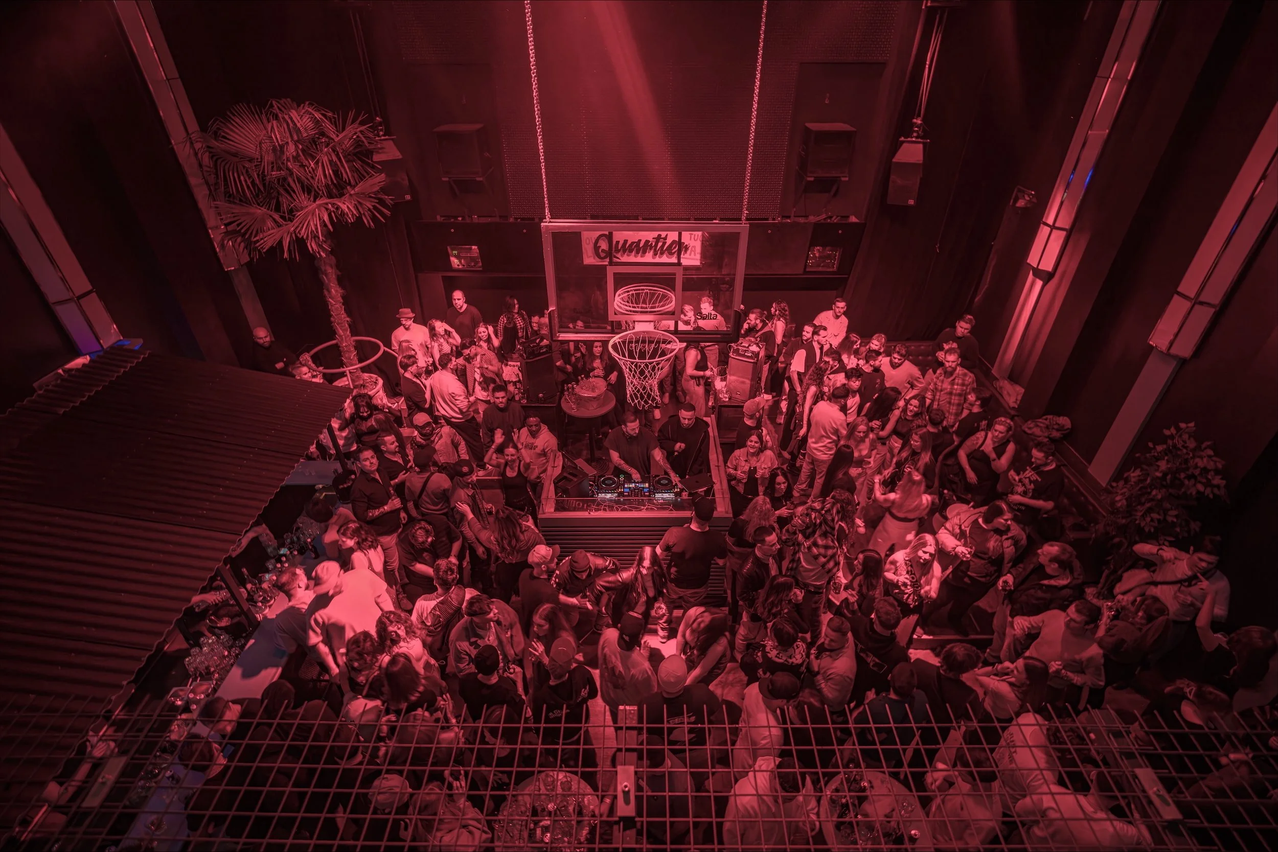 Menschen tanzen und feiern in einer Diskothek mit roter Beleuchtung, einem DJ am Mischpult, einer Basketball-Korbanlage im Hintergrund und Palmen auf der linken Seite.
