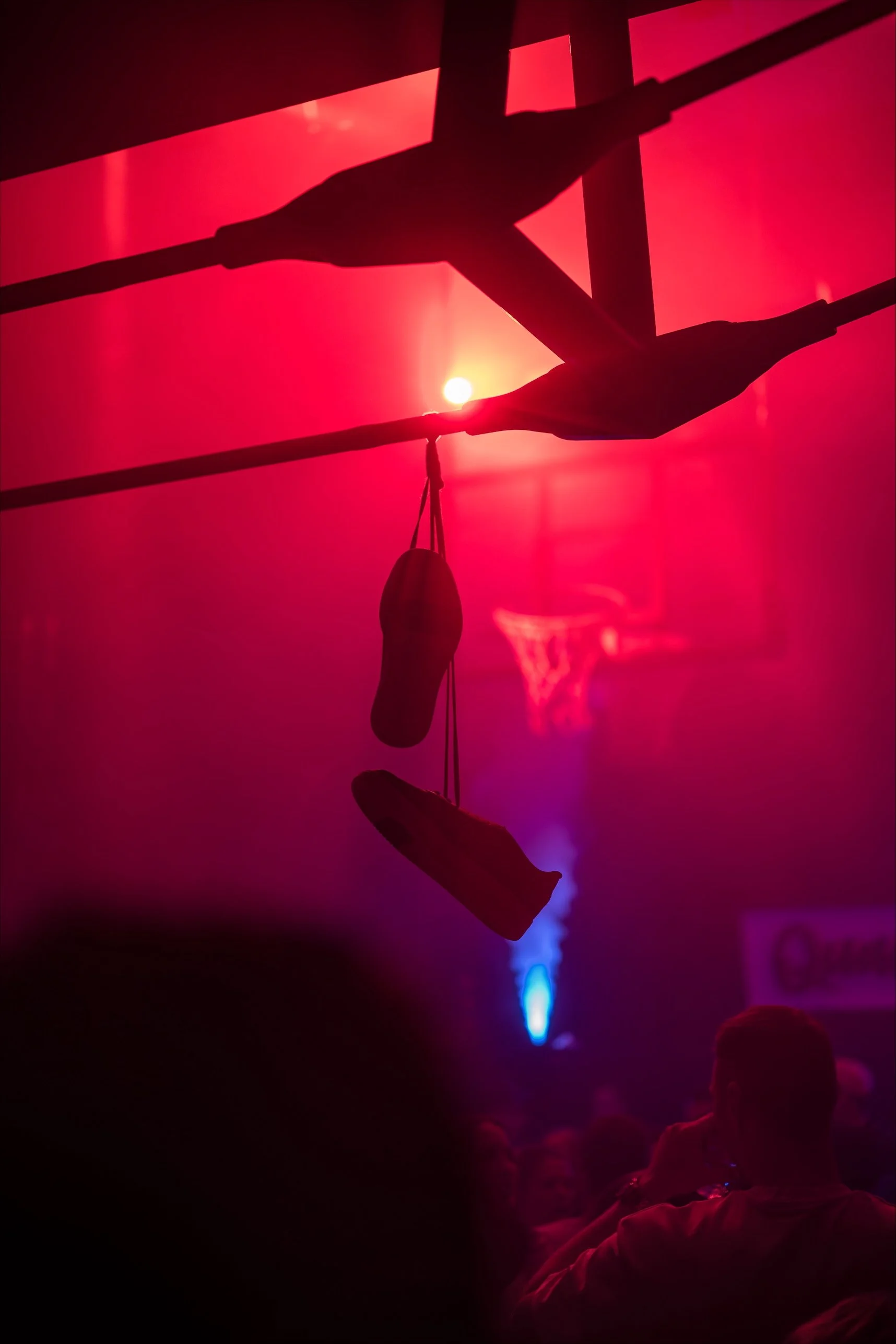 Dunkle Szene mit roten Lichtern, schwebenden Turnschuhen an Seilen, Menschen im Hintergrund und Basketballkörbe, Atmosphäre wie in einer Party oder Feier.