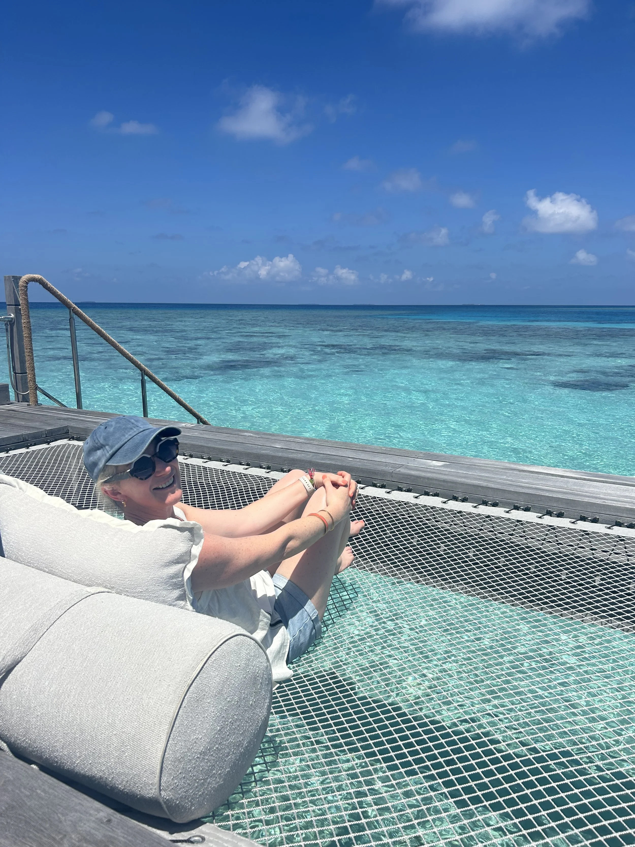Woman lying on a hammock on a wooden dock over clear turquoise ocean, wearing sunglasses, hat, white shirt, and shorts, smiling, with a bright blue sky and scattered clouds.