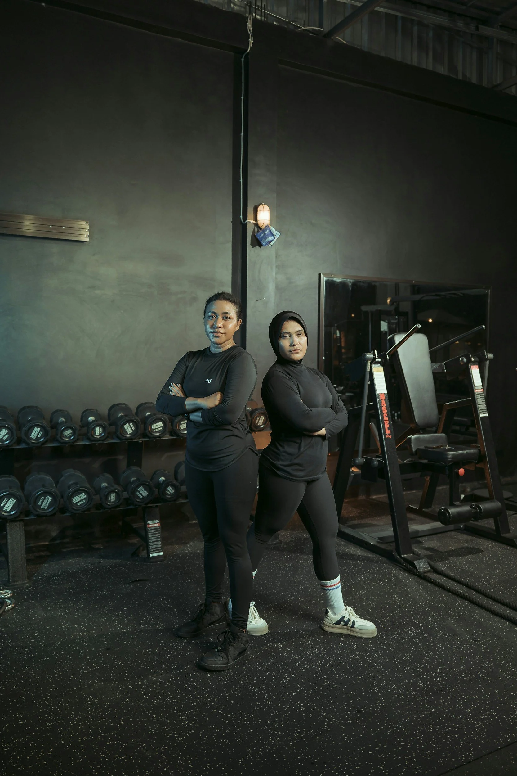 Two women in black athletic clothing standing with crossed arms in a gym, with dumbbells on racks and fitness equipment around them.