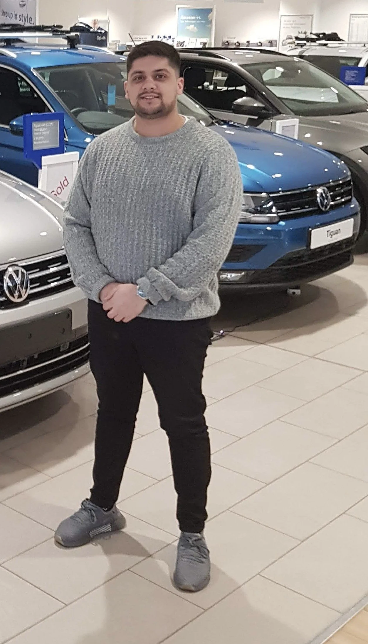 A man standing in a Volkswagen car dealership showroom with cars behind him, including a blue Volkswagen Tiguan.