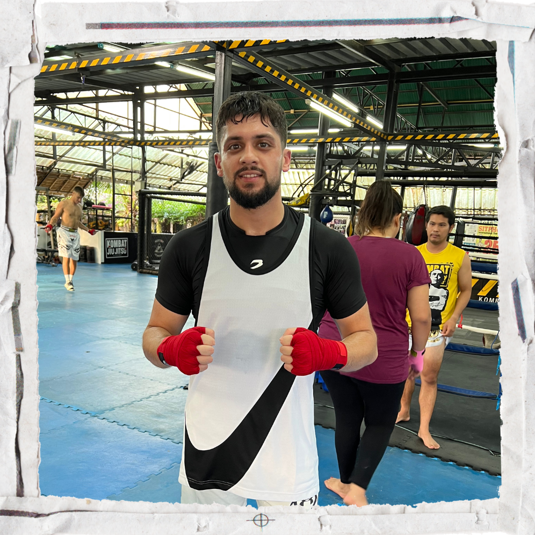A boxer in a gym wearing a white and black sports outfit and red hand wraps, standing in a fighting stance and smiling at the camera. In the background, there are training equipment, a person practicing kicks, and others training.