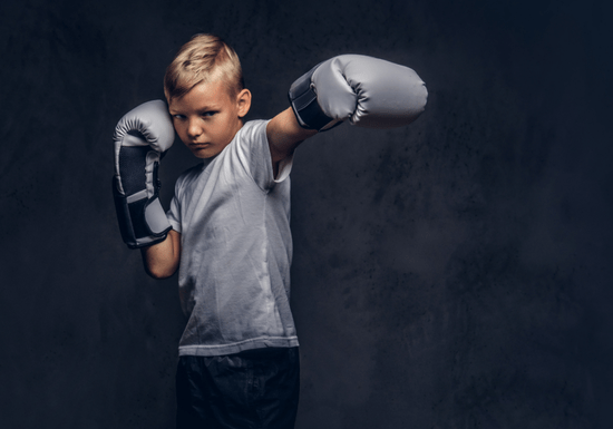 Young boy wearing boxing gloves practicing a boxing stance against a dark background.