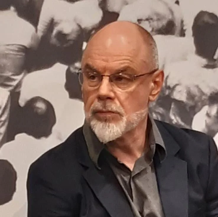 An elderly man with a bald head and glasses, wearing a dark blazer and shirt, looking to his left, in front of a black and white photograph of multiple people.