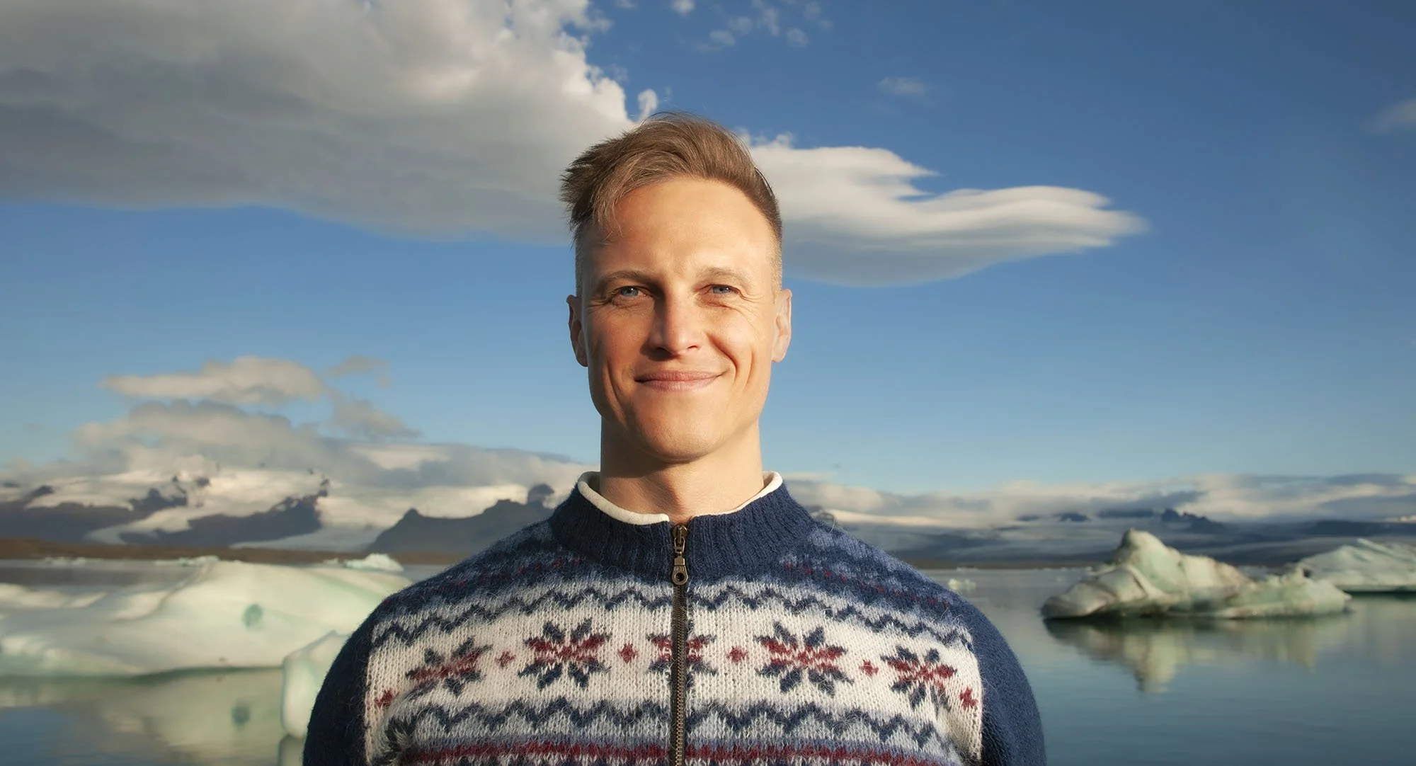 A young man smiling outdoors in front of a body of water with icebergs and a partly cloudy sky.