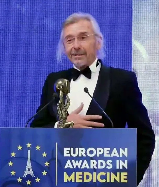 A man in a tuxedo with long gray hair and glasses speaking at a podium during the European Awards in Medicine event. The podium displays a graphic of the Eiffel Tower and the European Union flag.