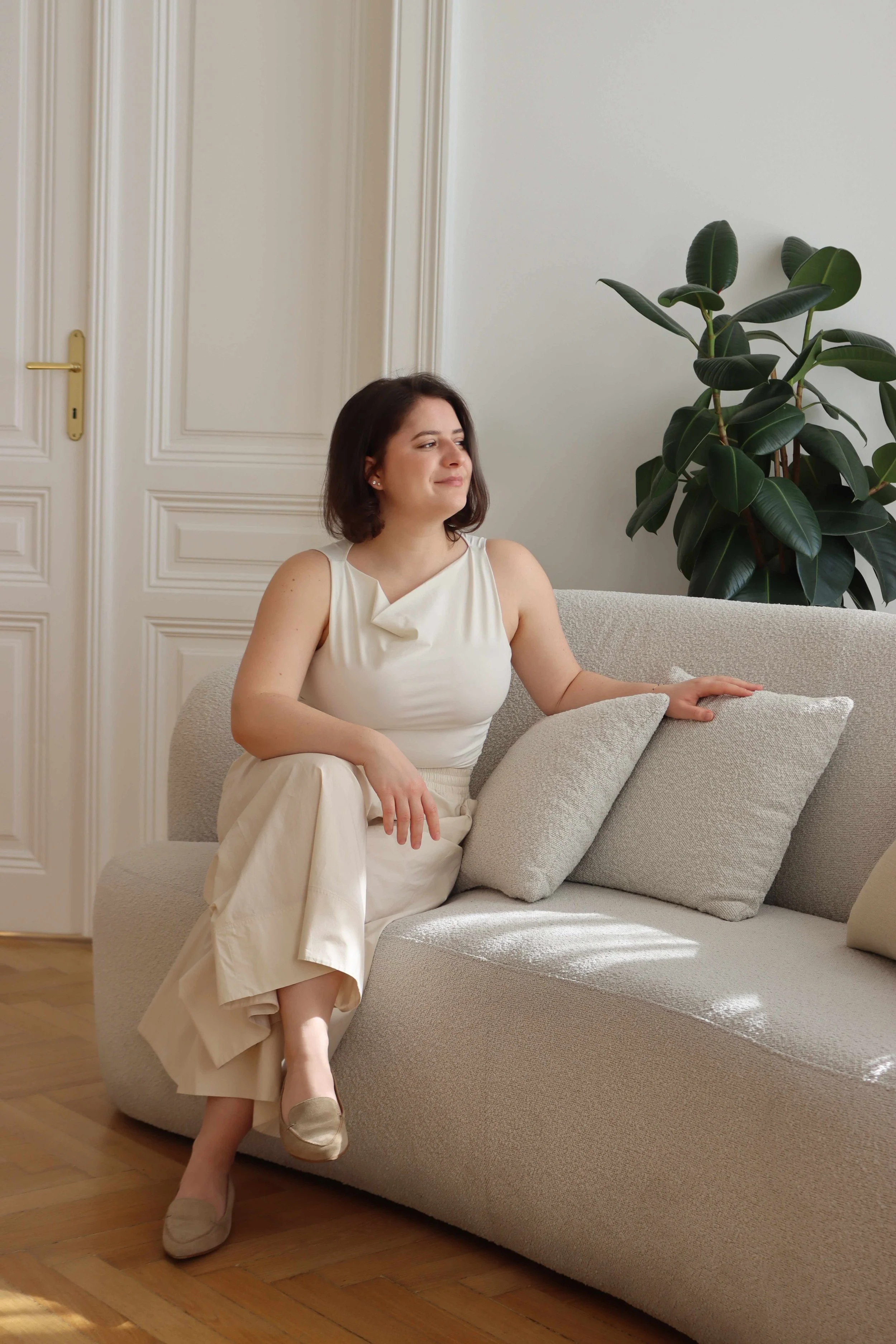 Junge Frau sitzt auf einem hellen Sofa in einem hellen Raum, neben einer großen Zimmerpflanze in einer modernen Altbauwohnung.