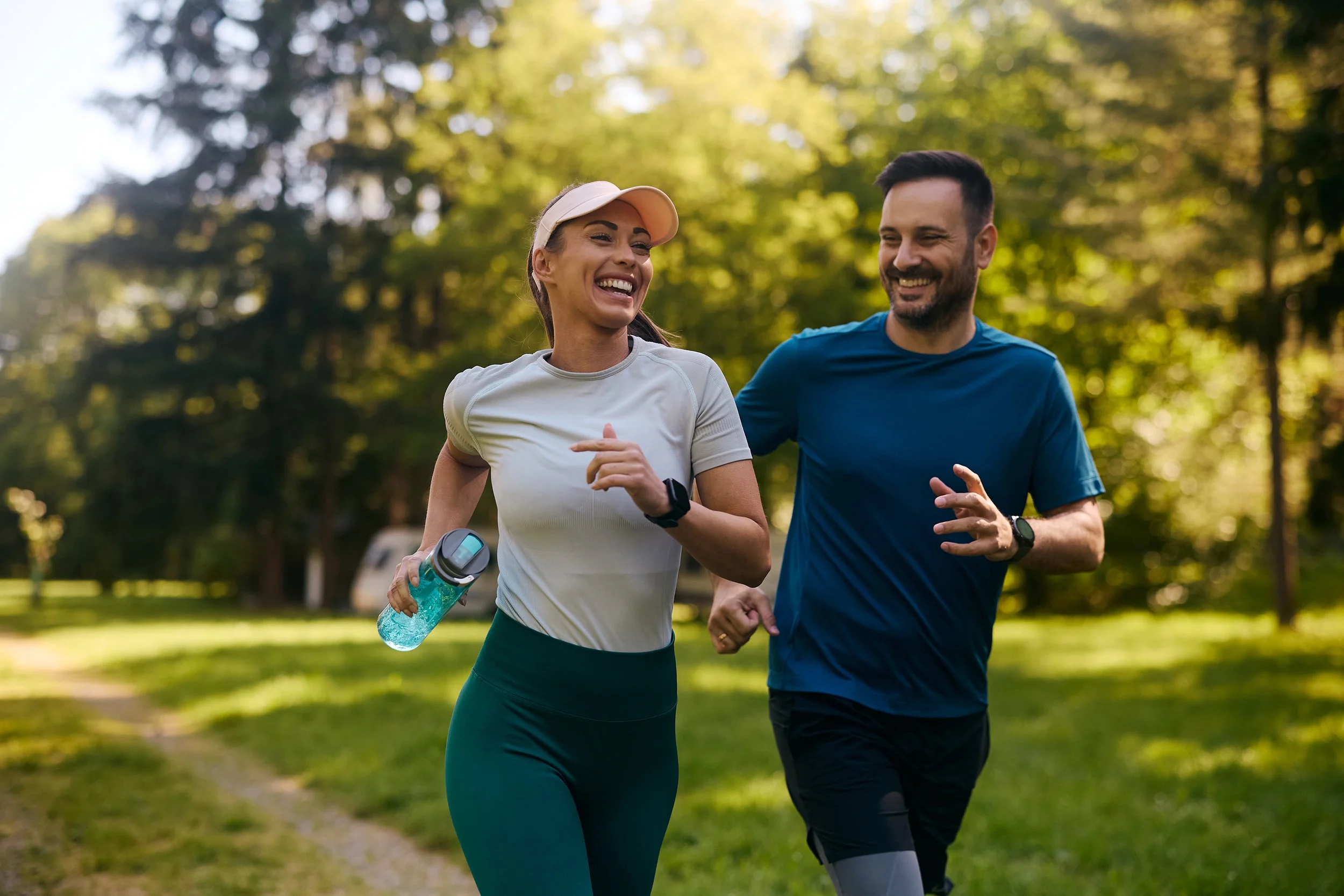 A man and woman are jogging outdoors in a park with trees in the background, smiling and enjoying their run.