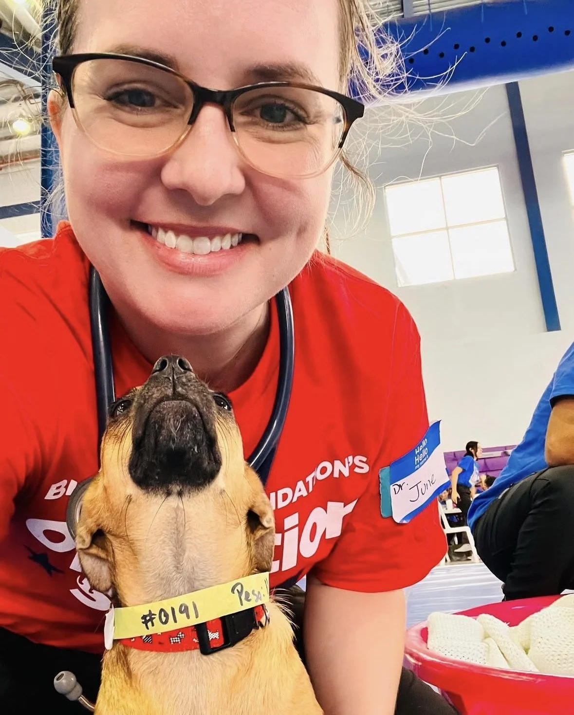 A smiling woman wearing glasses and a red shirt with a stethoscope around her neck is taking a selfie with a small dog. The dog is wearing a yellow collar with writing on it and is looking upwards. The woman has a name tag that says 'Dr. Julie.' They are indoors, possibly at an event, with other people present in the background.