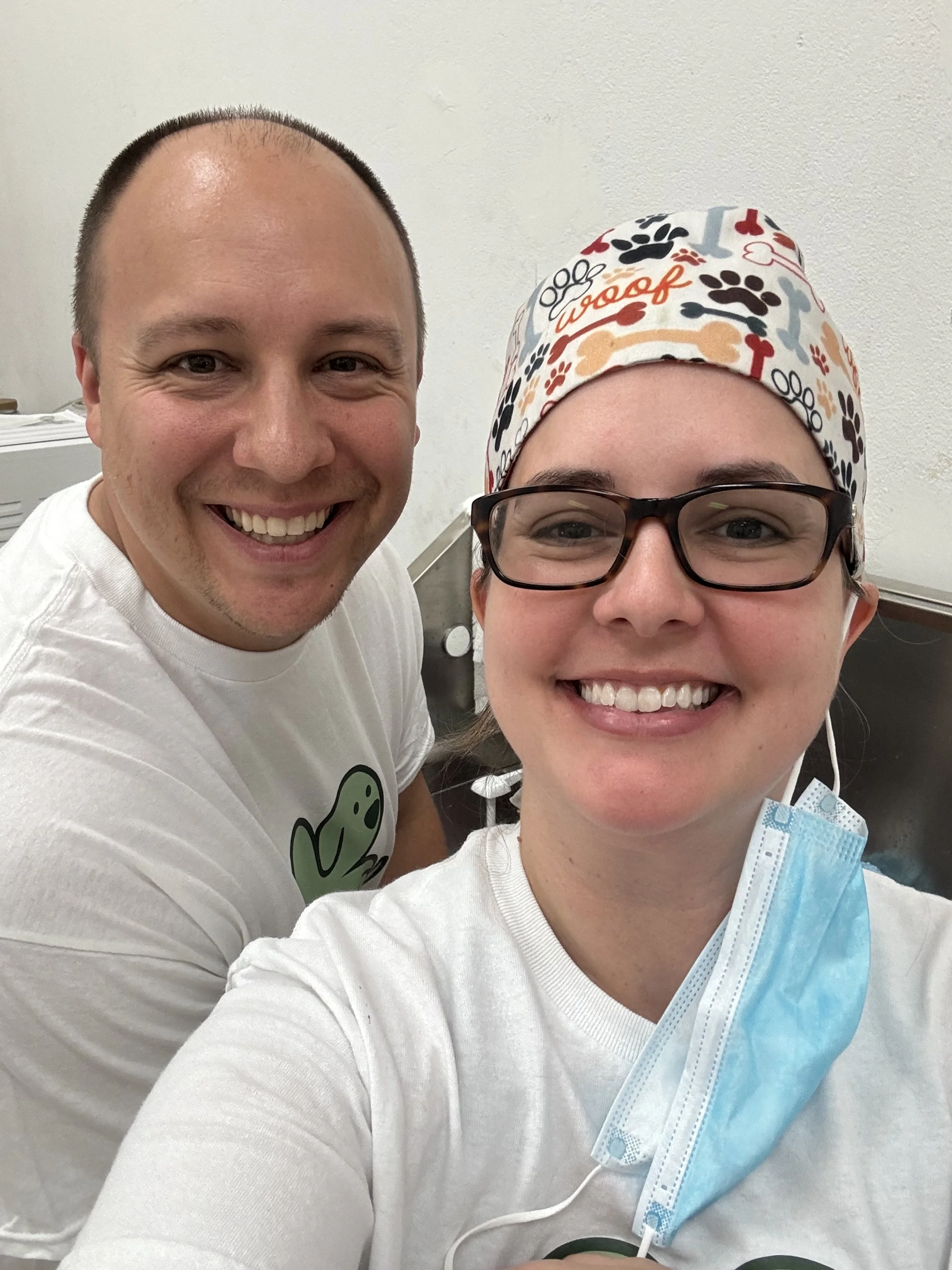 Two smiling healthcare workers, a man and a woman, taking a selfie in a clinical setting. The woman is wearing a colorful scrub cap, glasses, and a blue surgical mask hanging off her chin.