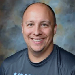 A smiling man with a shaved head wearing a dark Under Armour shirt, standing against a colorful, cloudy background.