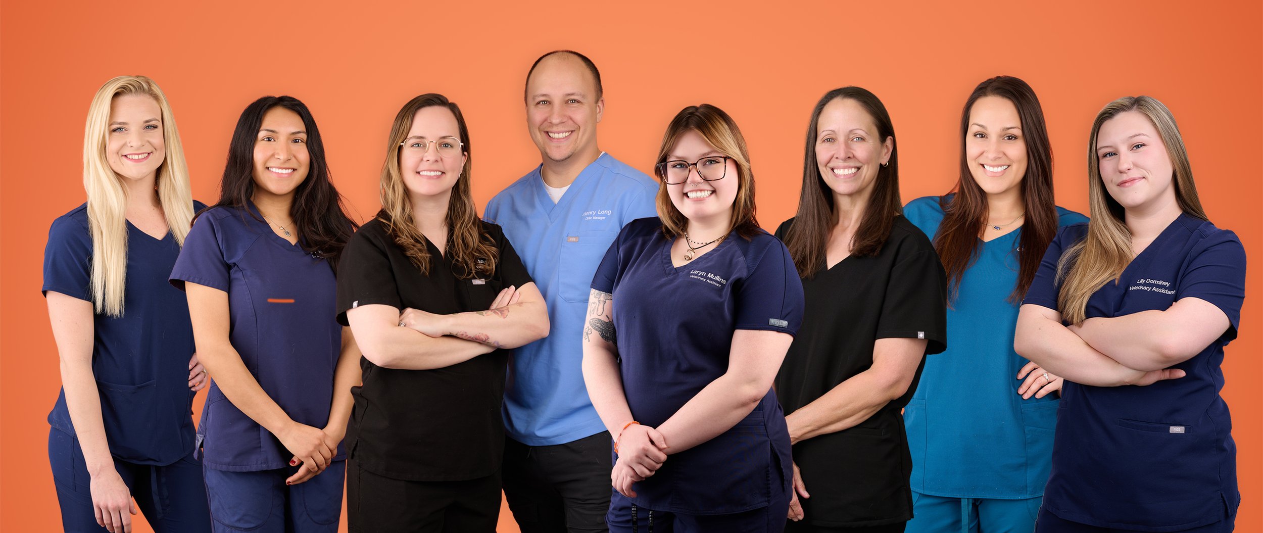 Group of healthcare professionals, mostly women, standing together against a warm orange background, smiling at the camera. They are wearing scrubs and lab coats.