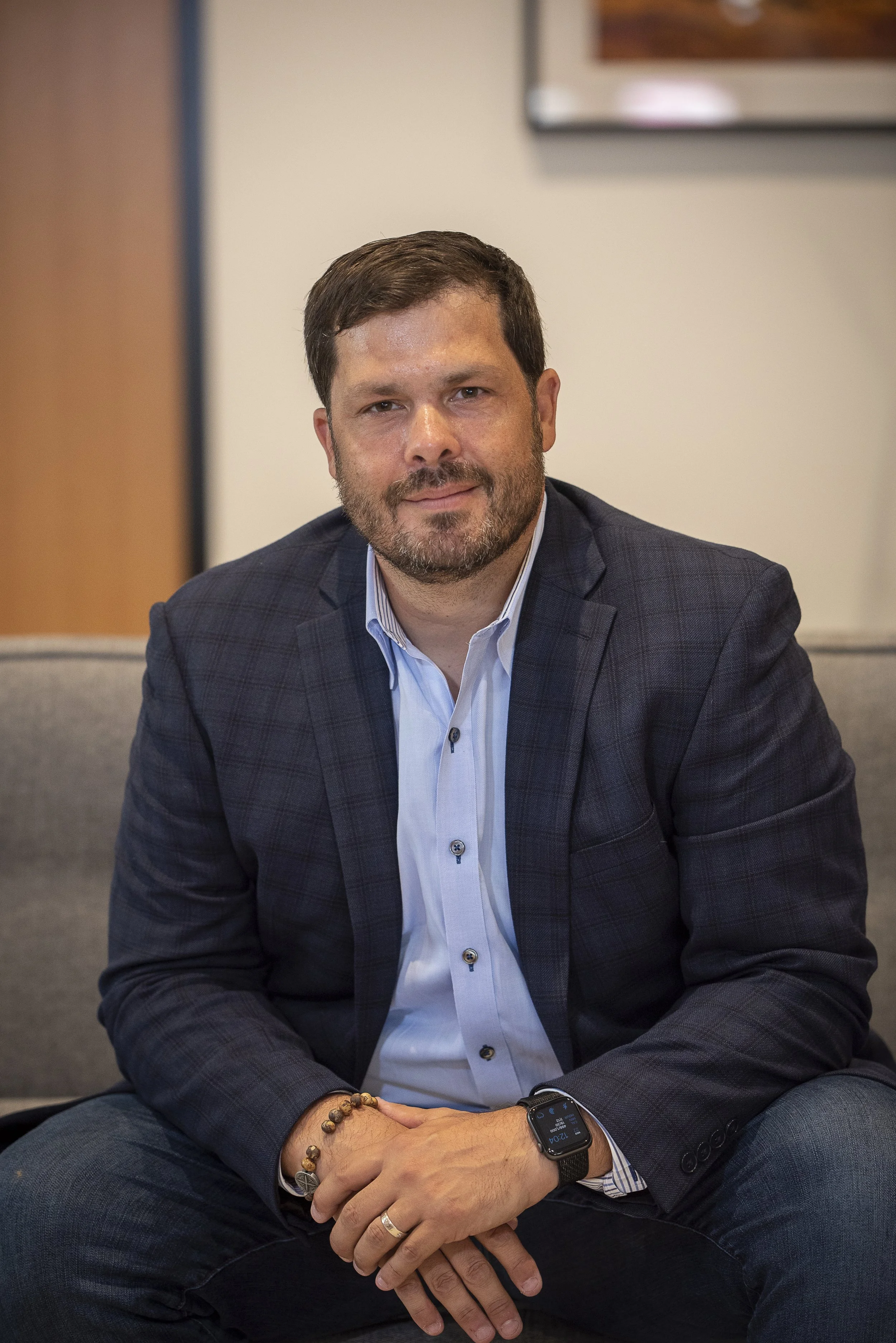 A man with short brown hair and a beard, wearing a dark checkered blazer and a white dress shirt, seated on a gray couch with his hands resting on his lap, in a professional indoor setting.