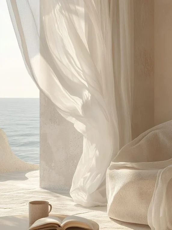 Sunlit interior space with flowing white curtains by a window overlooking the ocean, a beige sofa, an open book, and a coffee mug.