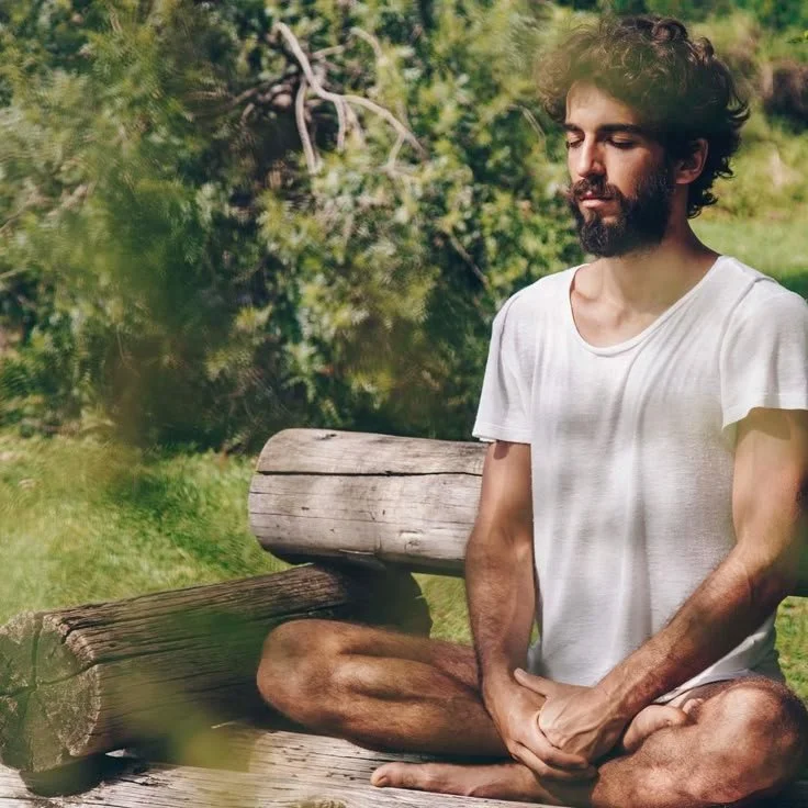 A man with a beard and mustache sitting cross-legged on a wooden bench outdoors, eyes closed, in a peaceful meditative pose amidst green trees and grass.