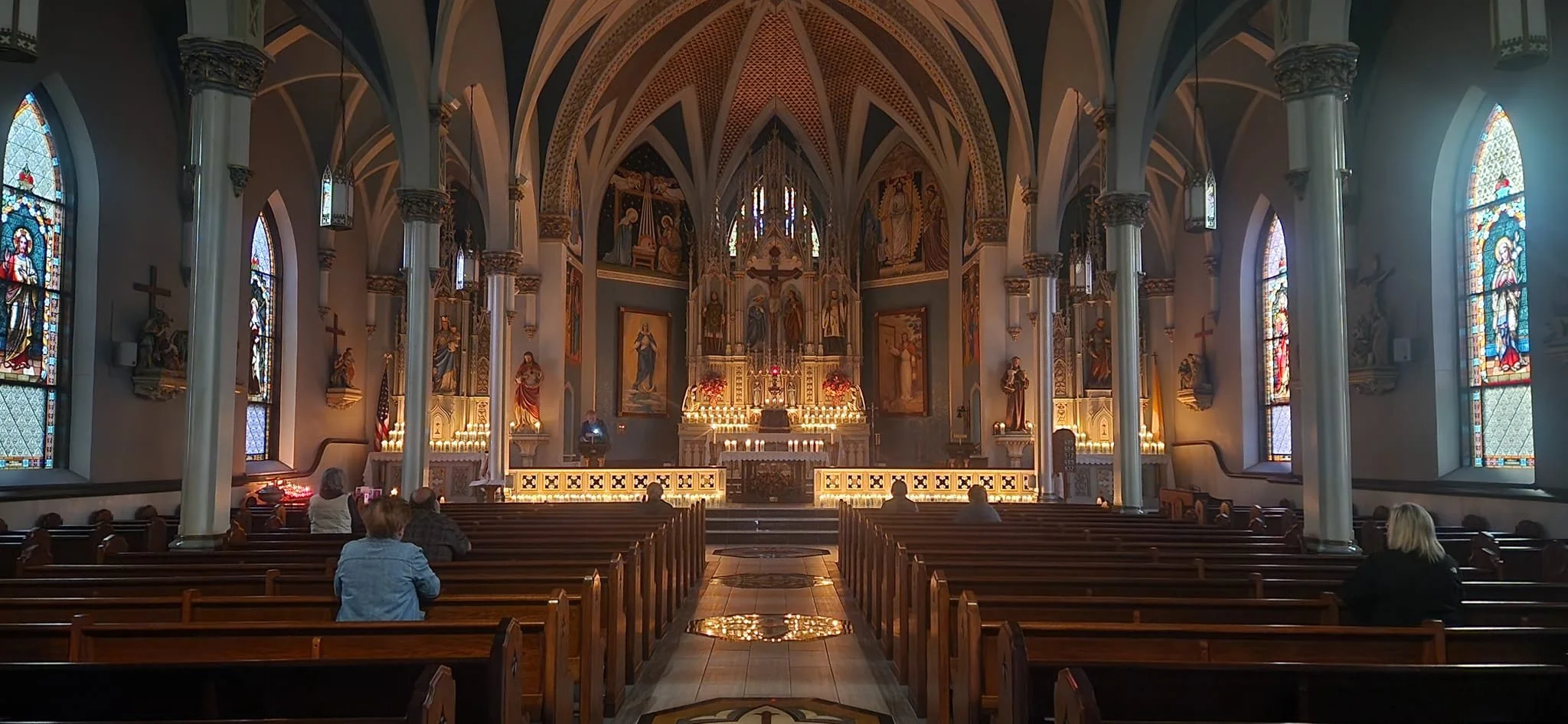 Interior of a church with high vaulted ceilings, stained glass windows, wooden pews, and an altar with candles and religious icons at the front.
