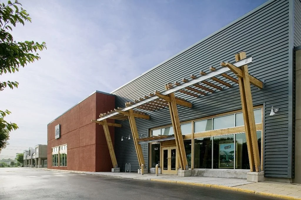 Exterior of a commercial building with modern architectural design, featuring large glass windows, a wooden pergola with angled supports above the entrance, and a gray and red facade.