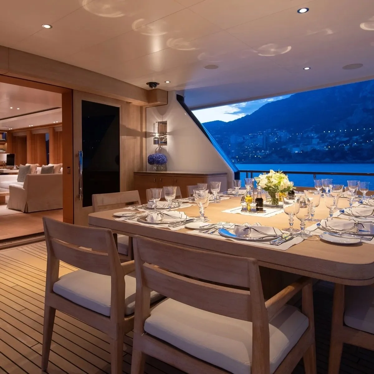 Elegant dining room with a large wooden table set for a meal, surrounded by wooden chairs with cushions, and decorated with a centerpiece of white flowers. The room has a view of a lake and mountains through large windows, with a cozy living area visible in the background, featuring white couches and pillows.