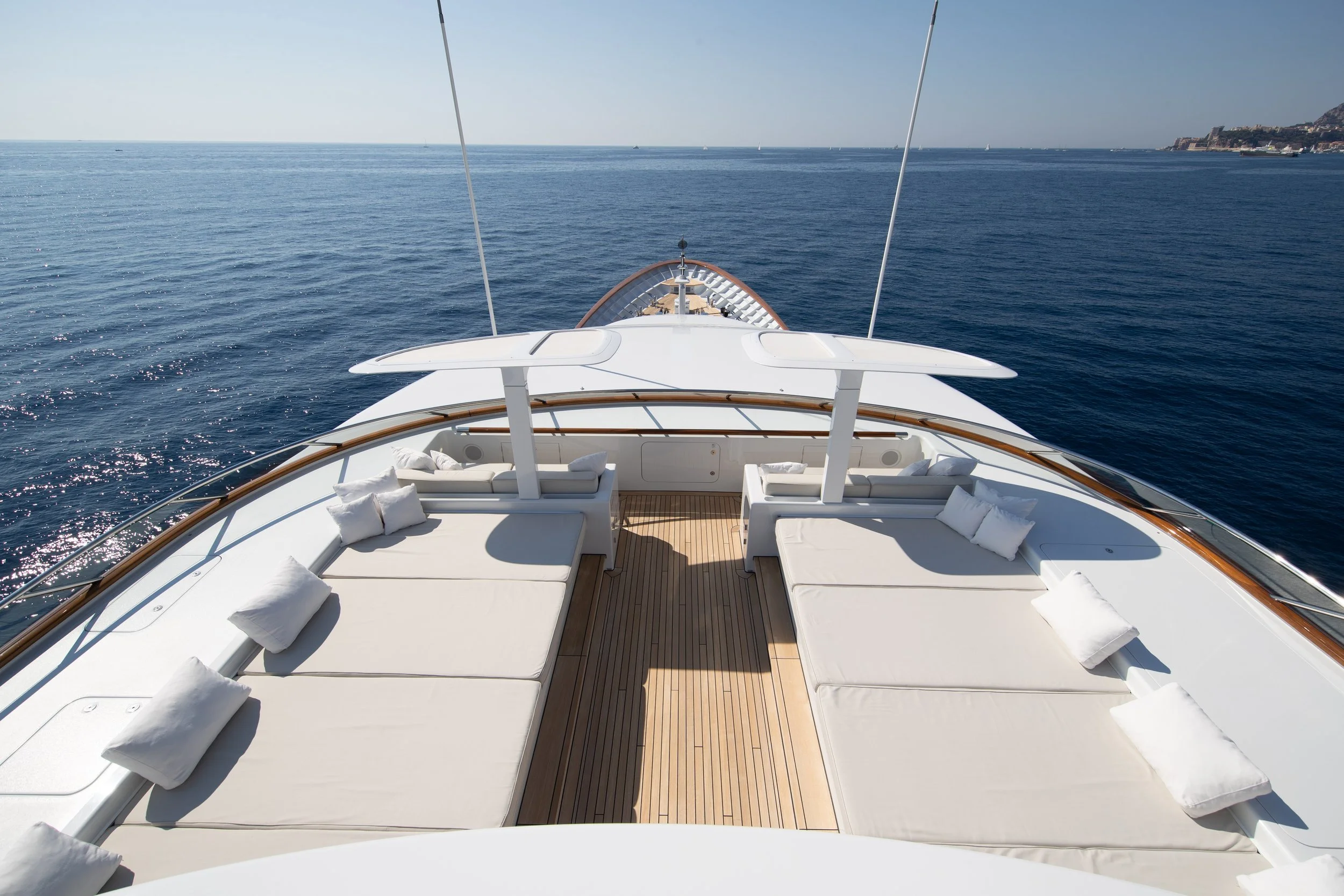 View from the deck of a luxurious yacht with many cushioned sunbeds, overlooking calm blue sea with small sailboats in the distance and a distant coastline with hills.