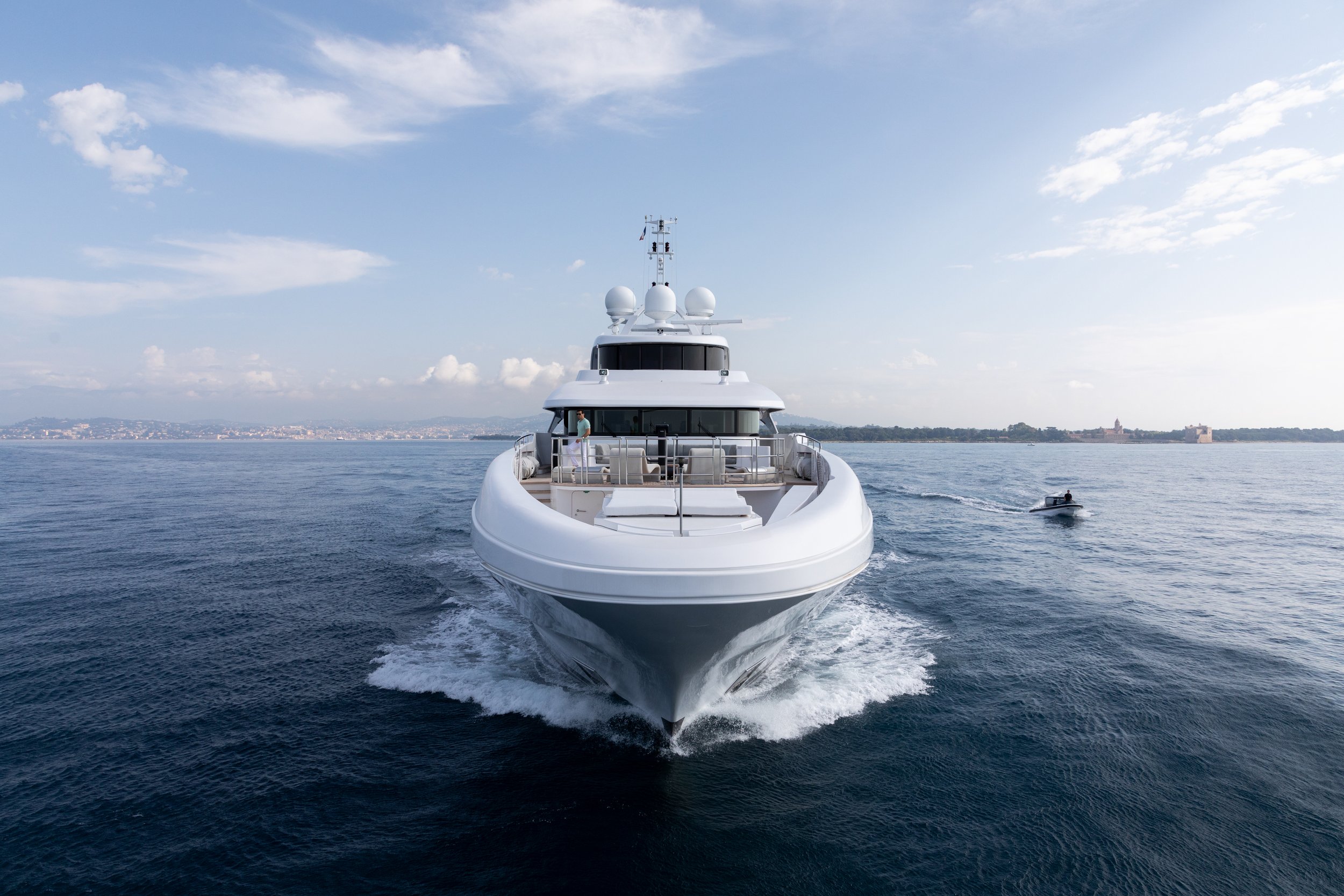 Large white yacht with radar and antennas on the deck sailing on calm ocean, with a small motorboat nearby and distant shoreline under a partly cloudy sky.
