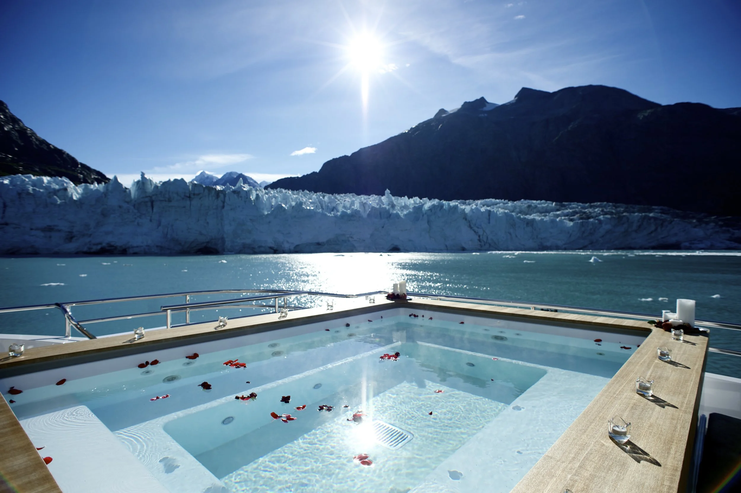 A luxurious hot tub with rose petals floating on the water, set on a boat with a view of a glacier, mountains, and a bright sun in a cold, icy landscape.