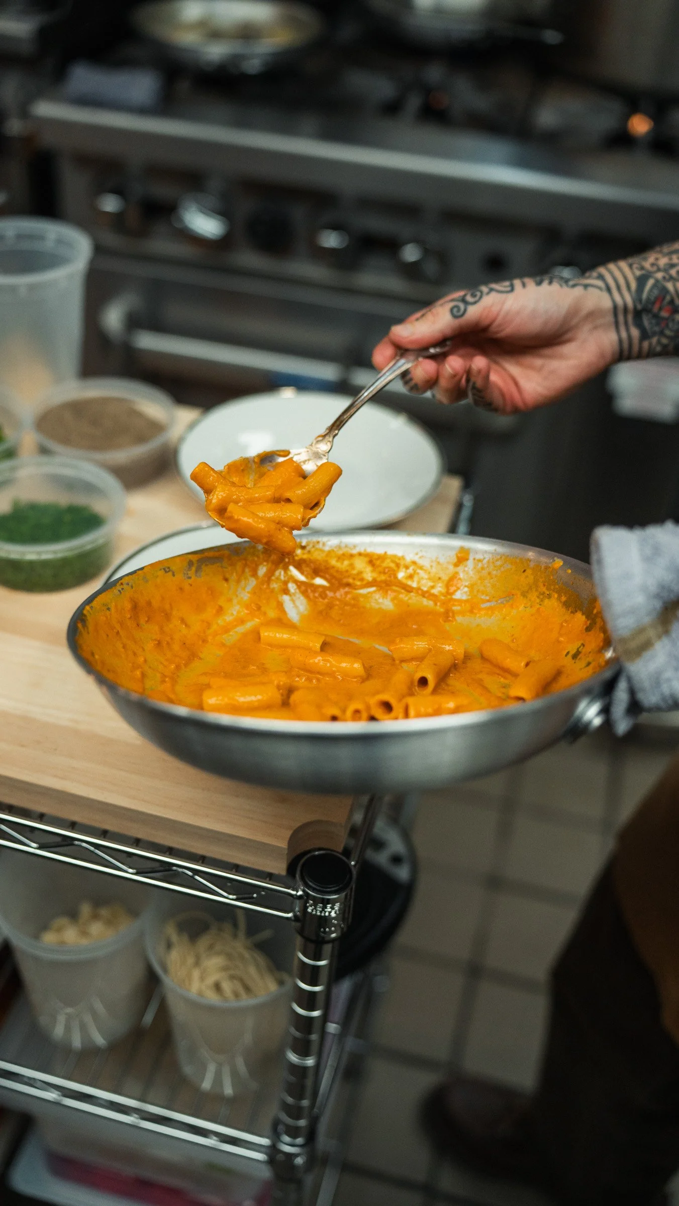 Person scooping orange tomato-based pasta onto a fork from a metal bowl with cooked pasta in a kitchen.