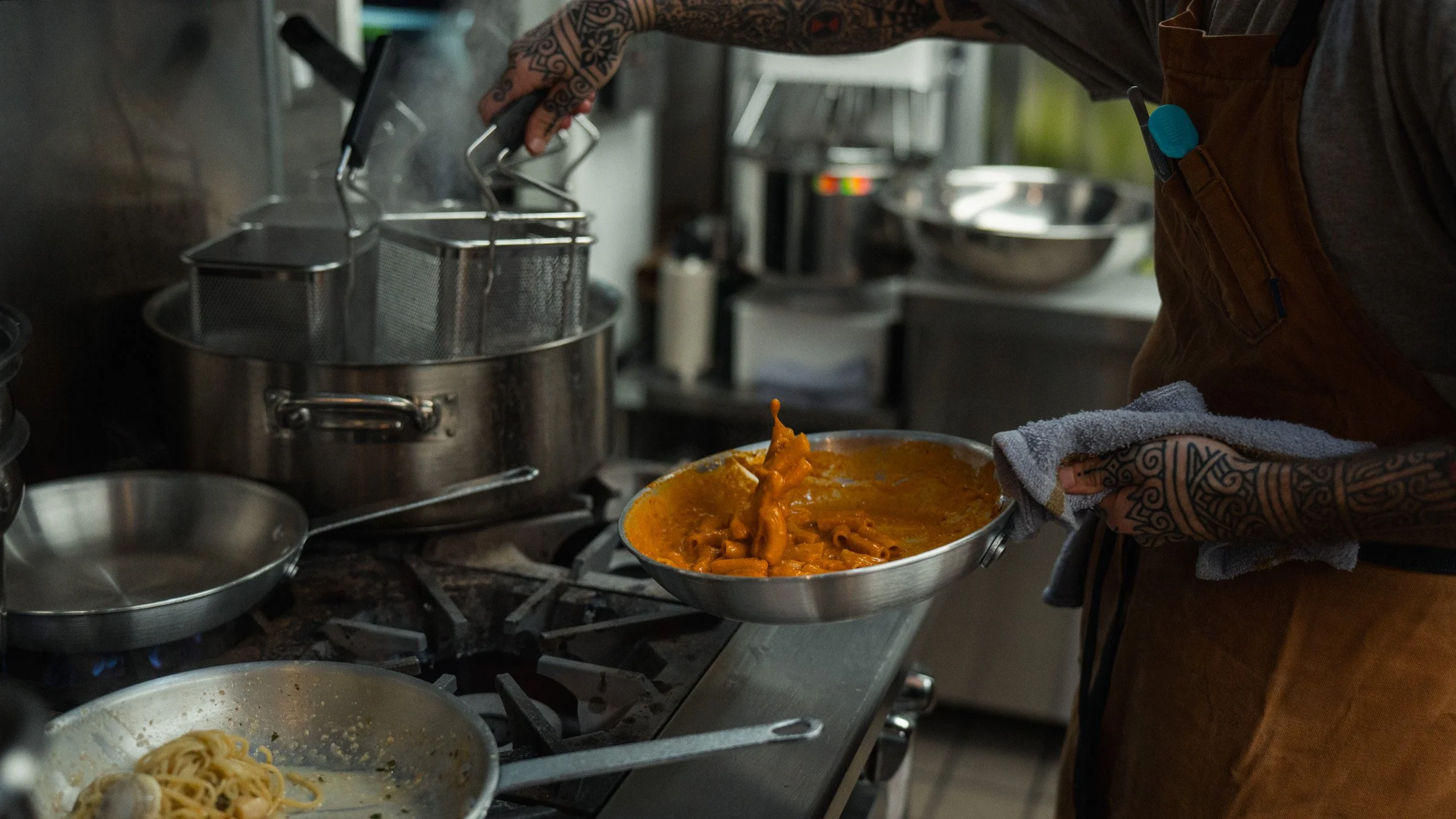 A person with tattoos on their arm is cooking pasta with tomato sauce in a kitchen. They are holding a pan with pasta and sauce using a towel, and appear to be stirring or serving it. There are various kitchen utensils and containers around, includin