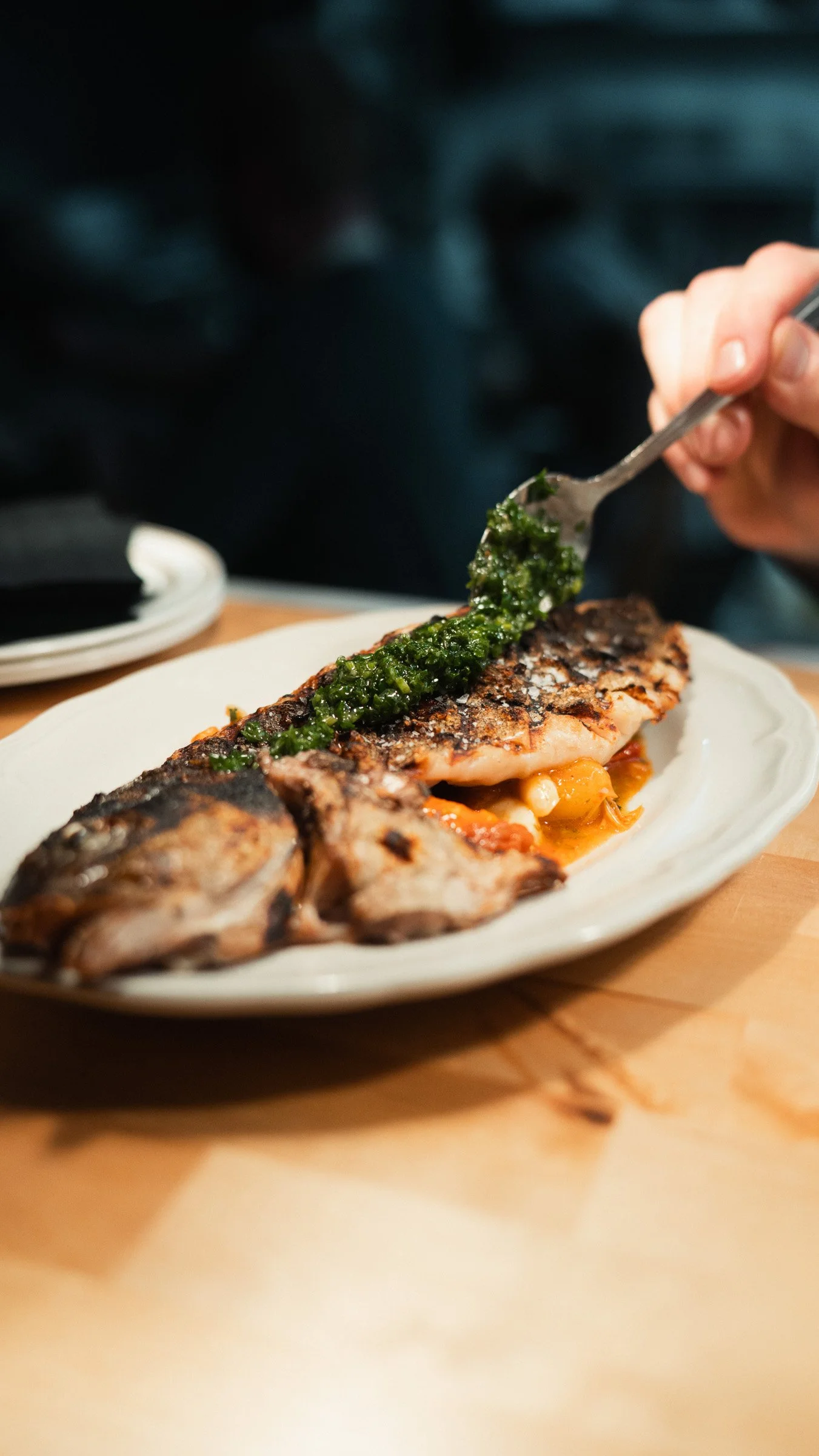 A person adding green sauce to a grilled fish fillet on a white plate. The plate contains roasted vegetables underneath the fish, and the person is holding a fork with the green sauce.