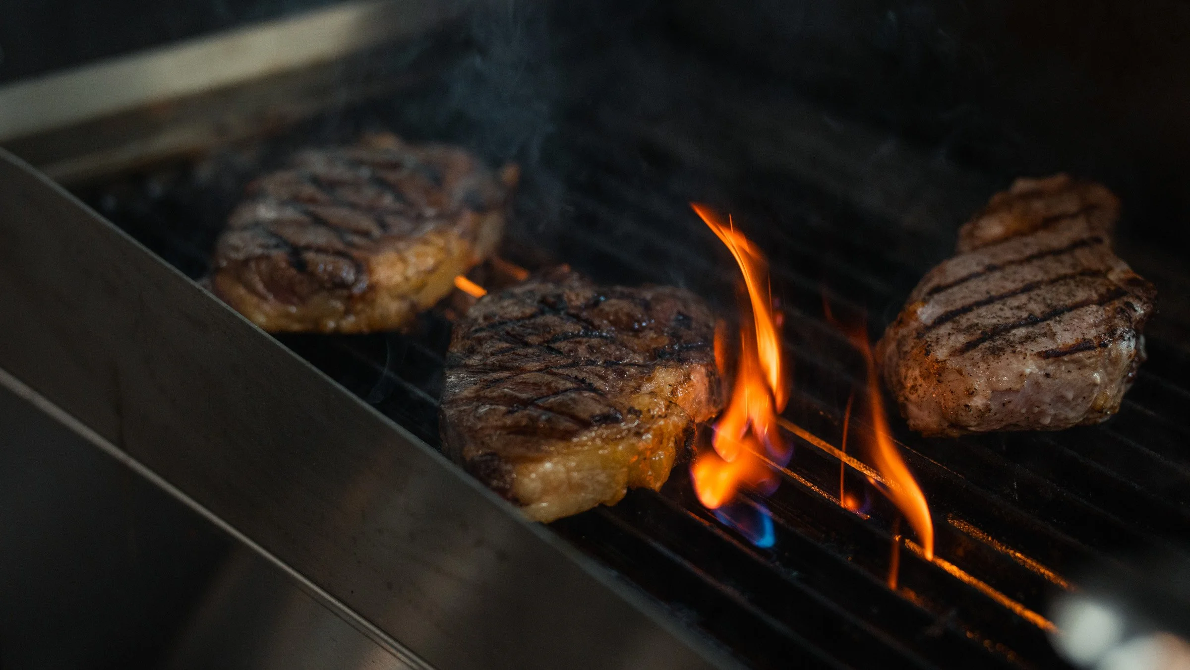 Three pieces of seasoned steak grilling on a barbecue with flames and grill marks visible.