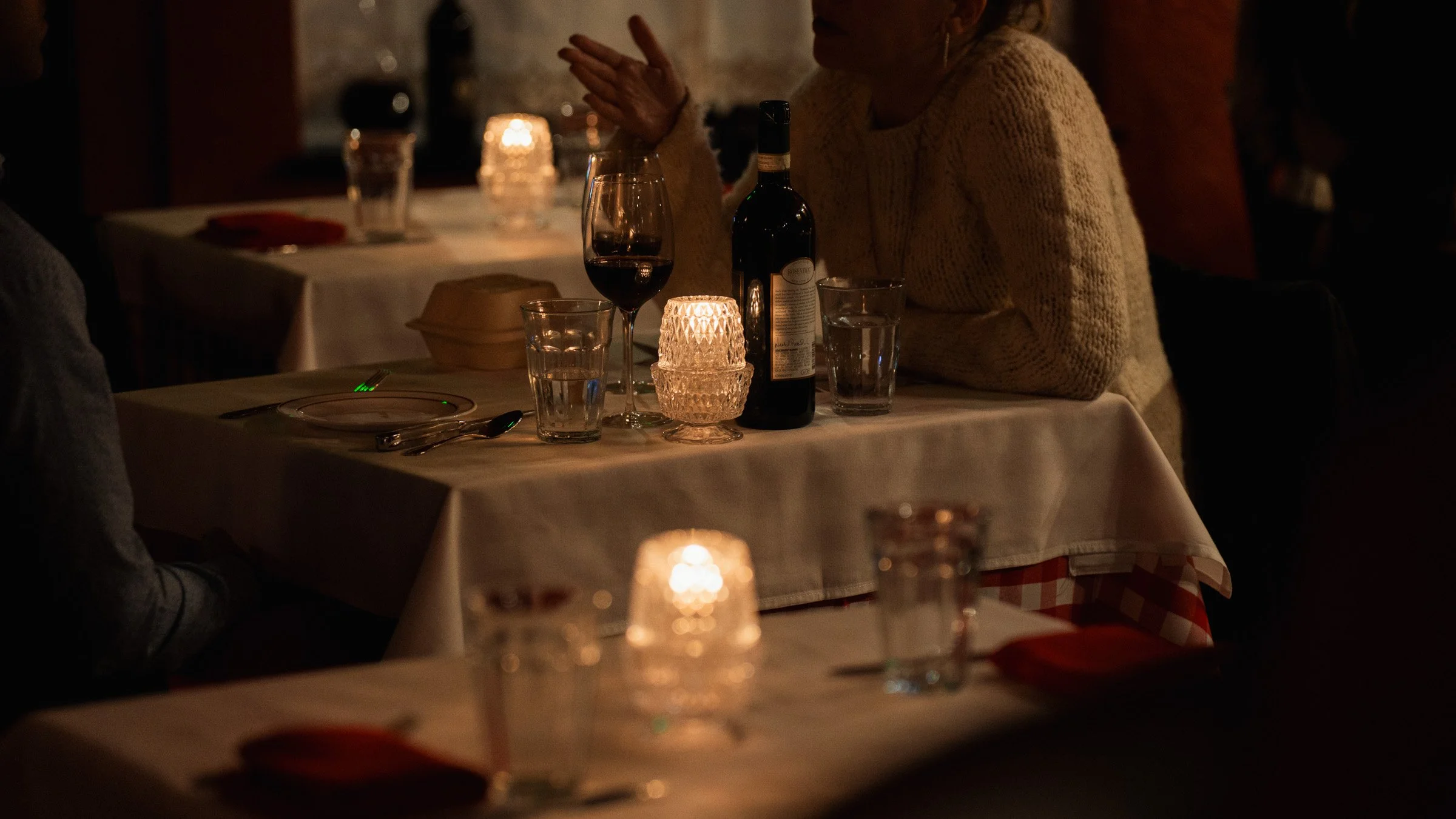 A dimly lit restaurant table set with a wine glass, a bottle of wine, and candles, with a person wearing a beige sweater sitting at the table.