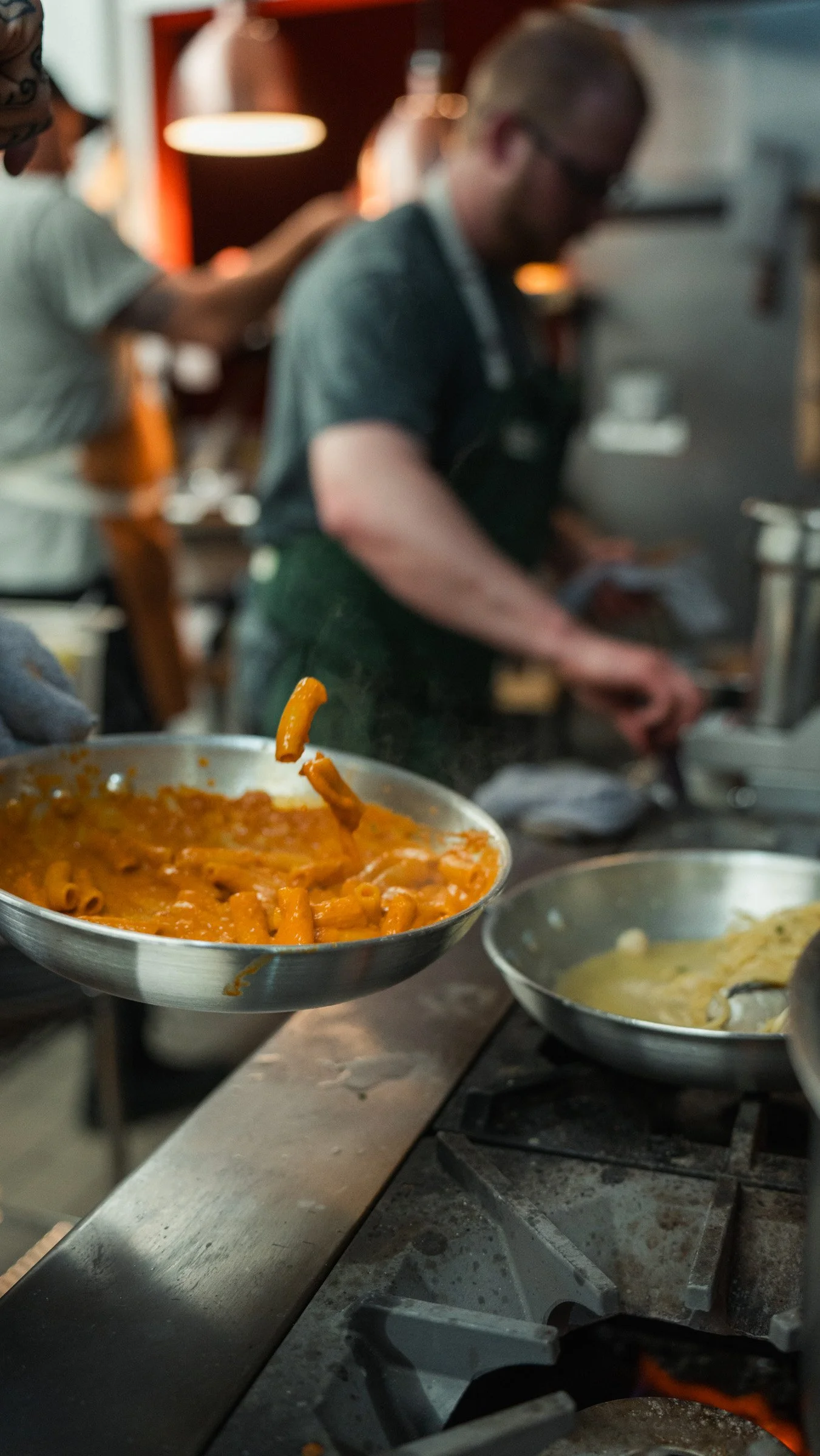 A chef throwing pasta with tomato sauce from a pan in a professional kitchen, with another dish nearby.