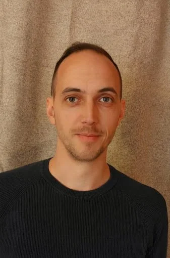 Portrait d'un homme avec une peau claire, des cheveux courts et une expression neutre, portant un pull noir devant un fond en tissu beige.