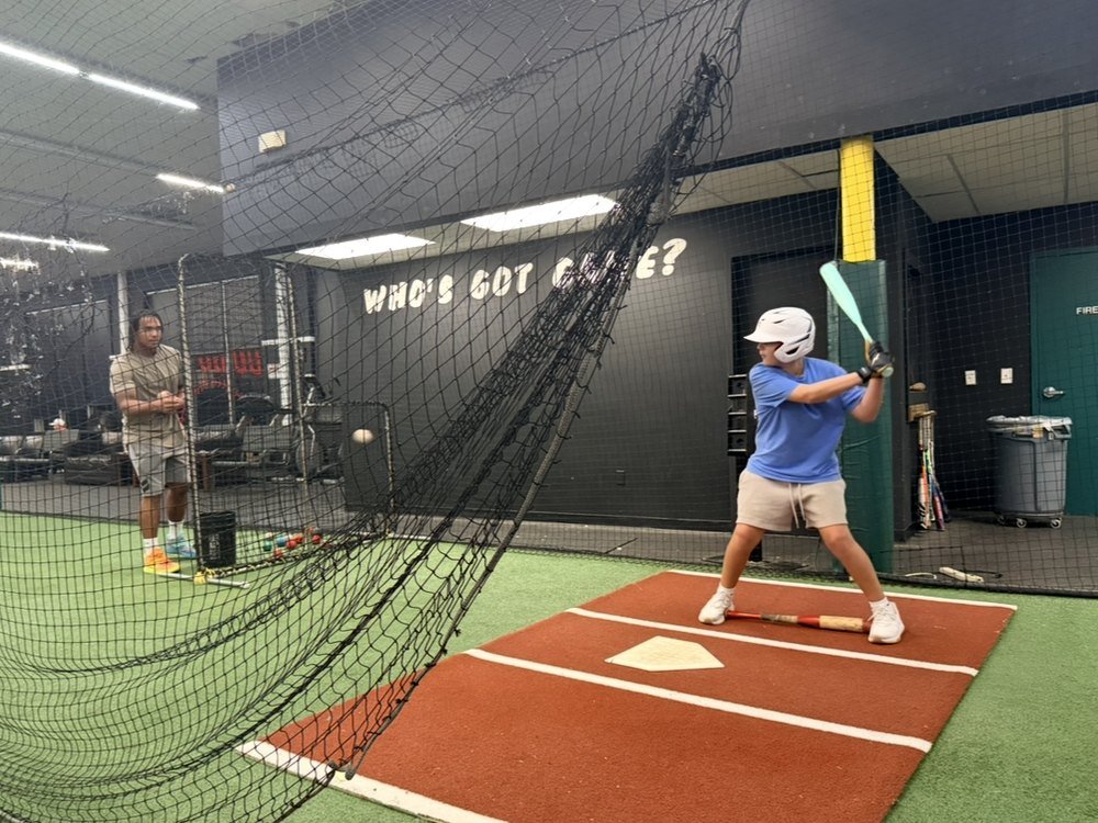 An MFIG hitter in the batting cage taking a swing while a coach watches