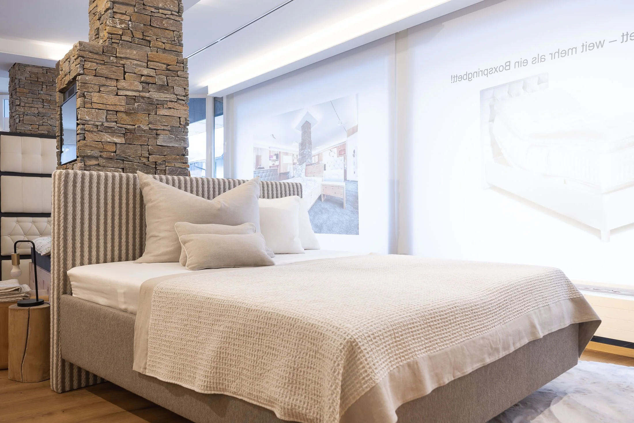 Schlafzimmer mit neutraler Farbgebung, Bettrahmen mit Stoffbezug, Kissen und Decke, Fenster mit weißen Vorhängen, Wanddekoration mit Bildern, Steinsäule und Holzdecke.
