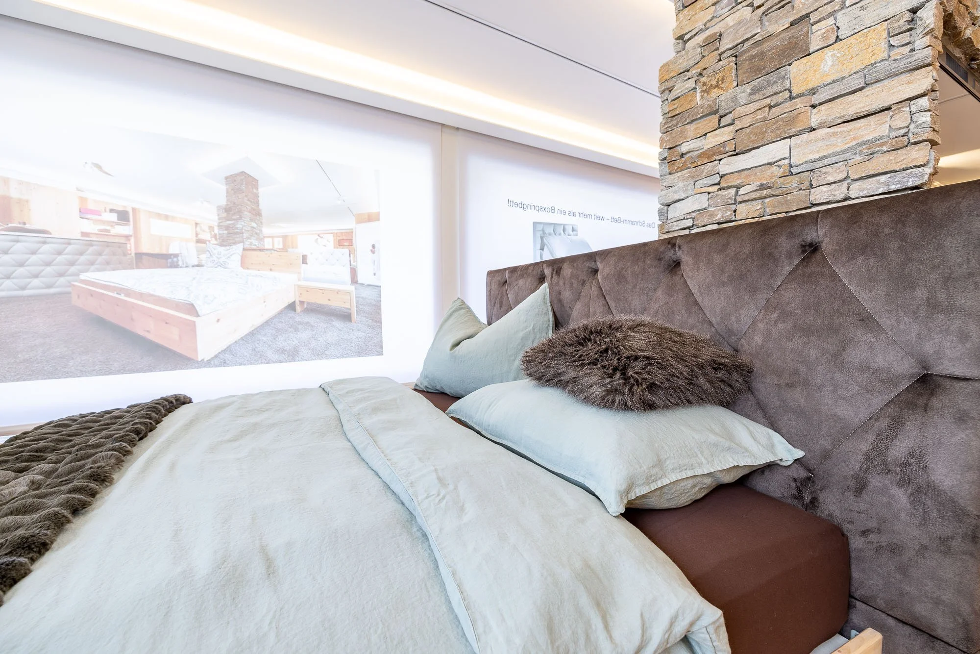 Bett im Schlafzimmer mit grauem Kopfteil, hellen Bettwäsche, einer pelzigen Kissen, und einem Blendenbildschirm im Hintergrund. Die Wand ist teilweise mit Steinen verkleidet.