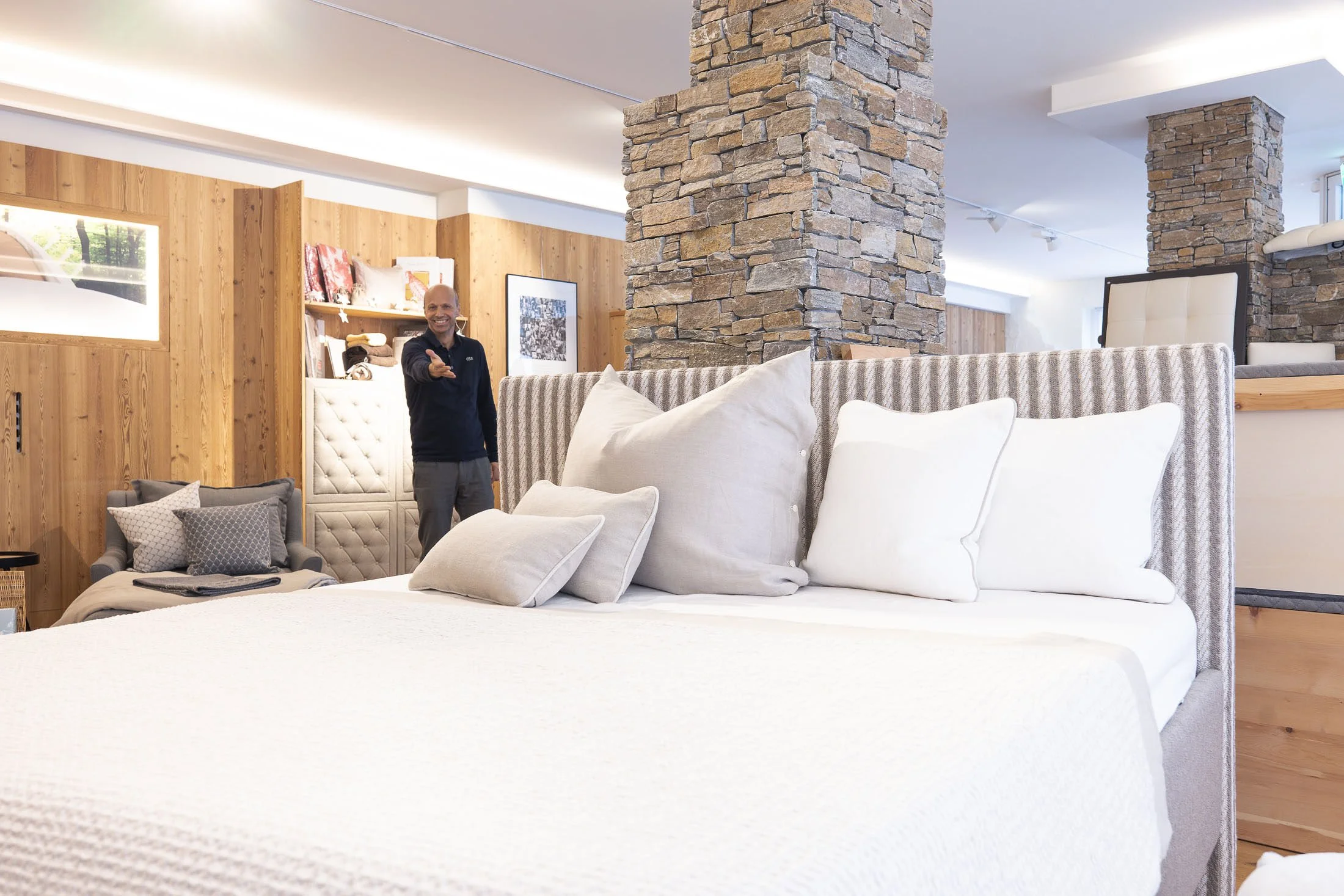 Ein Mann steht in einem modernen Schlafzimmer mit Holz-Wandpaneelen, grauen Möbeln und einem gemauerten Kamin, der Bett ist mit vielen weißen und grauen Kissen dekoriert.