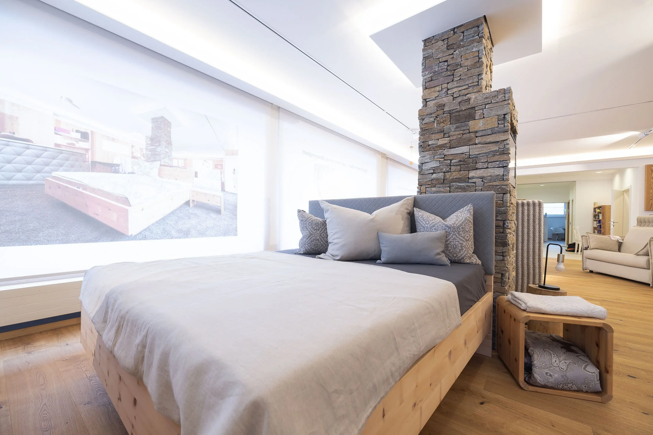 Ein modernes Schlafzimmer mit Holzboden, hellen Wänden und einem Bett mit grauem Kopfteil und mehreren Kissen. Es gibt eine Trennwand aus Naturstein und ein Fenster mit Vorhängen, durch das Tageslicht ins Zimmer fällt. Im Hintergrund sind weitere Woh