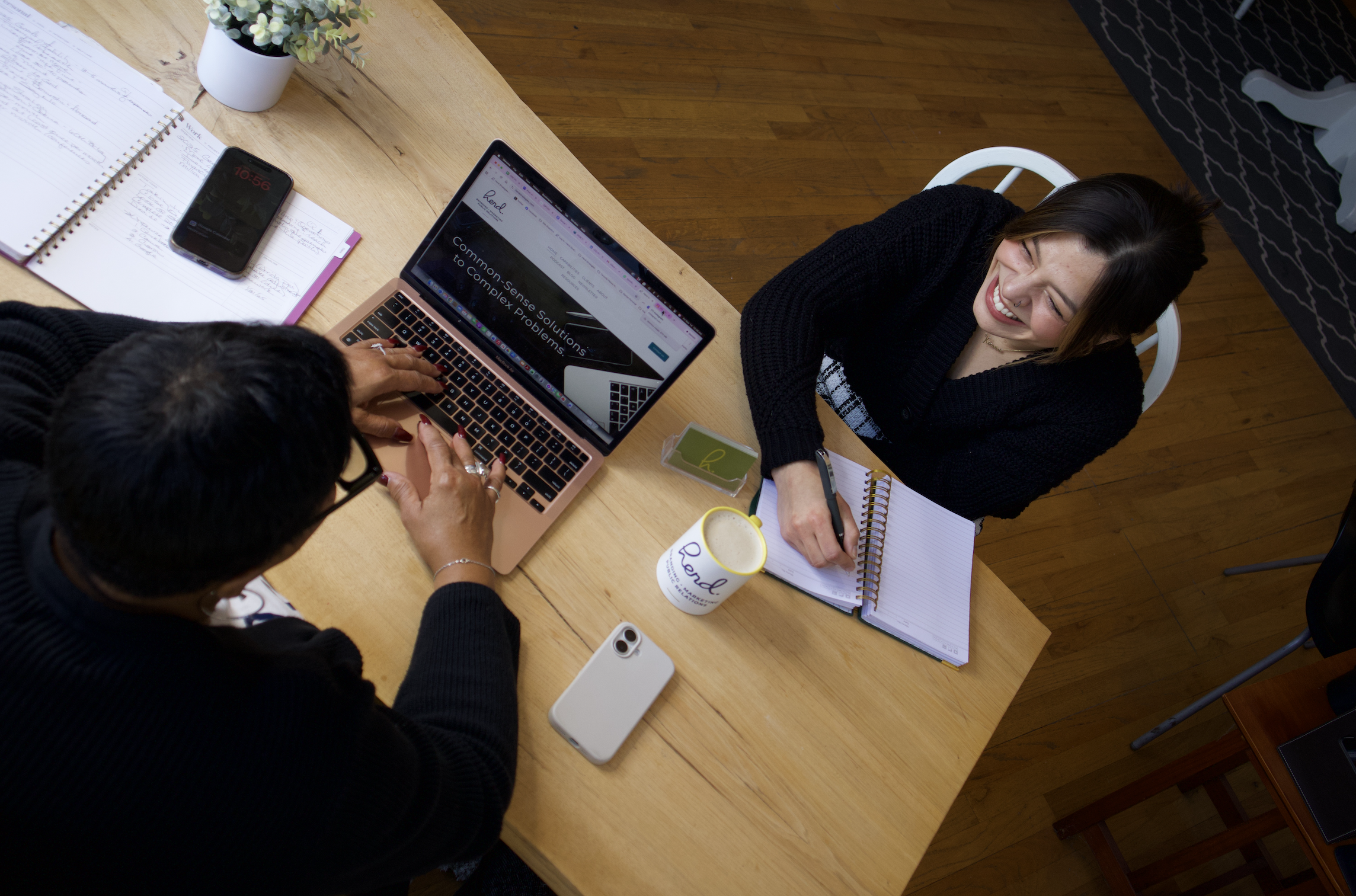 Two women having a meeting at a wooden table, with notebooks, a laptop, a smartphone, a cup, and a small box on the table. One woman is sitting and smiling, the other is working on the laptop.
