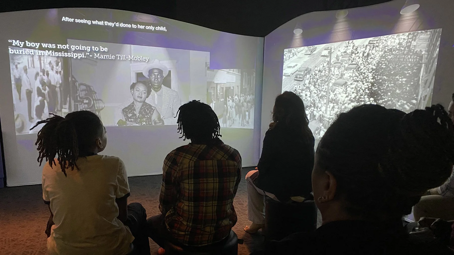 People sitting and watching a black-and-white documentary or presentation projected on a wall, featuring an image of a woman and a man with text displaying a quote from Mamie Till-Mobley.