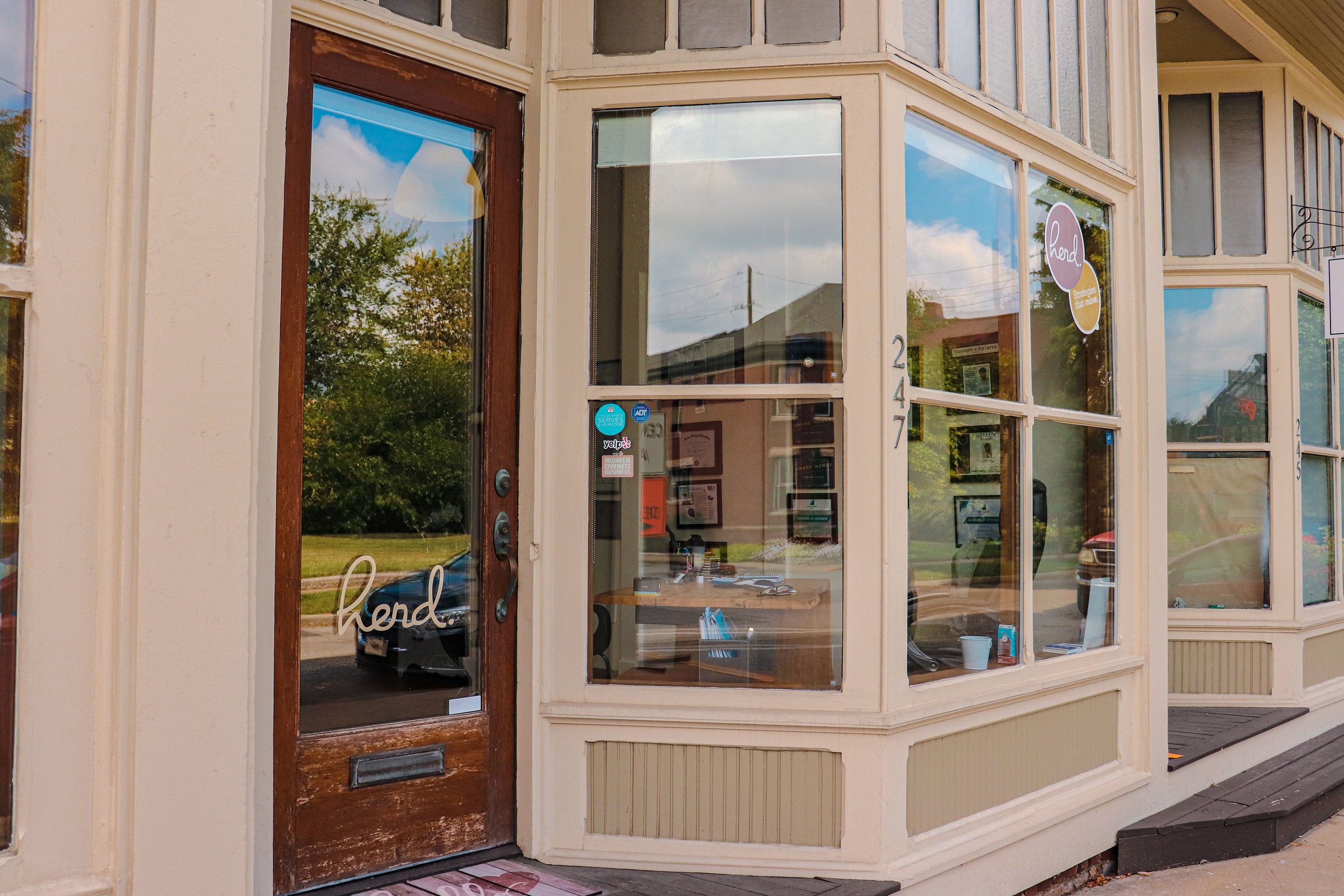 The storefront of a business at 247 with a glass door and large front windows, with the word 'hend' written on the door in cursive. Inside, there are framed certificates or pictures on the wall and a desk with office supplies.
