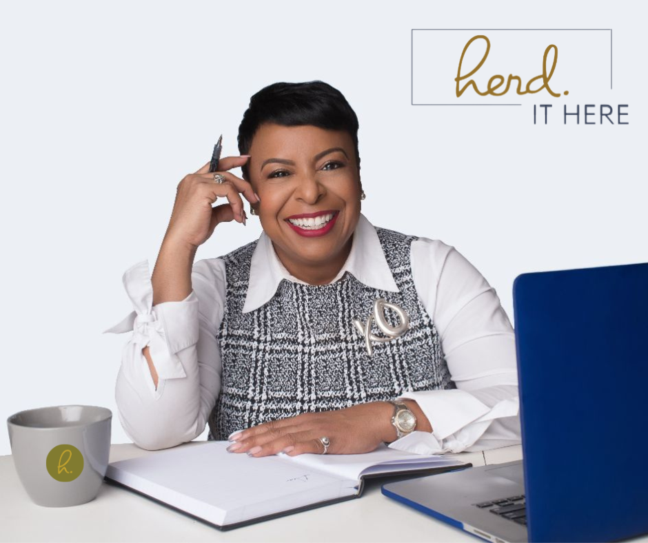 A smiling woman with short dark hair sitting at a desk, holding a pen to her temple, with a coffee mug, open notebook, and a blue laptop in front of her, and a sign that says "HERD IT HERE" on the wall.