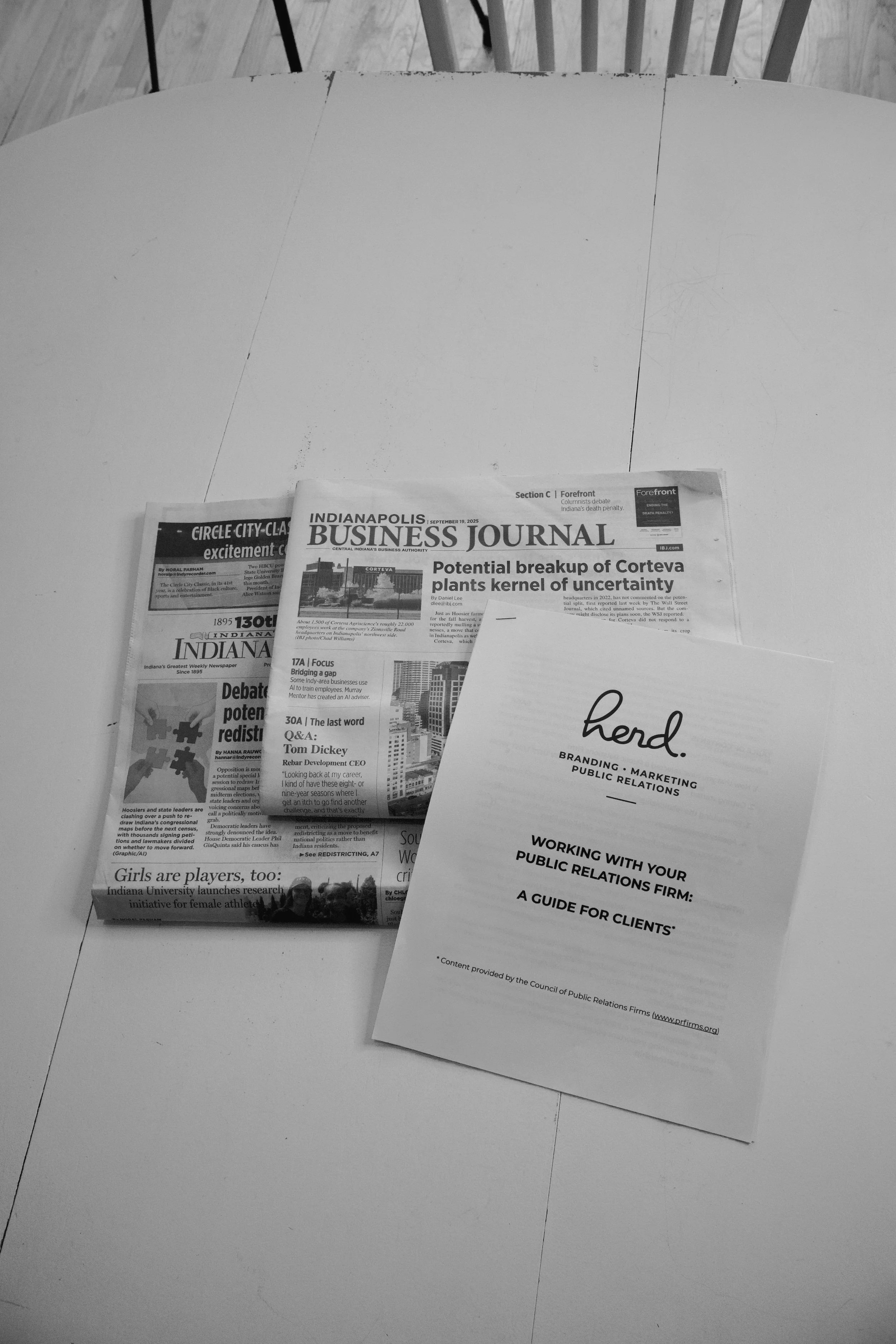 A white round table with a newspaper titled 'Indianapolis Business Journal' and a pamphlet titled 'herd' on top of it, with a wooden chair visible in the background.